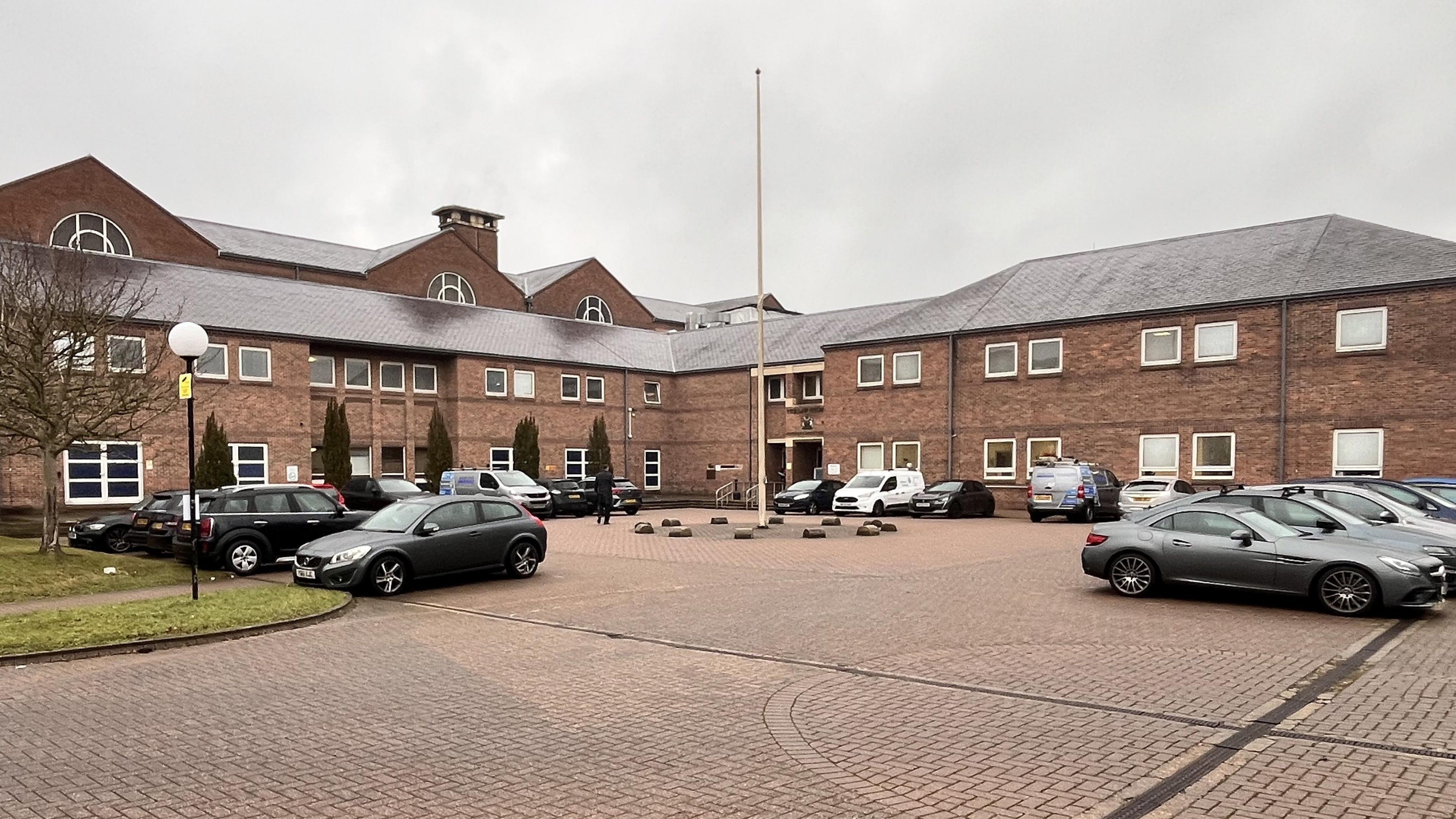 A two-storey L-shaped court building with a brick-paved forecourt and a number of cars parked outside.
