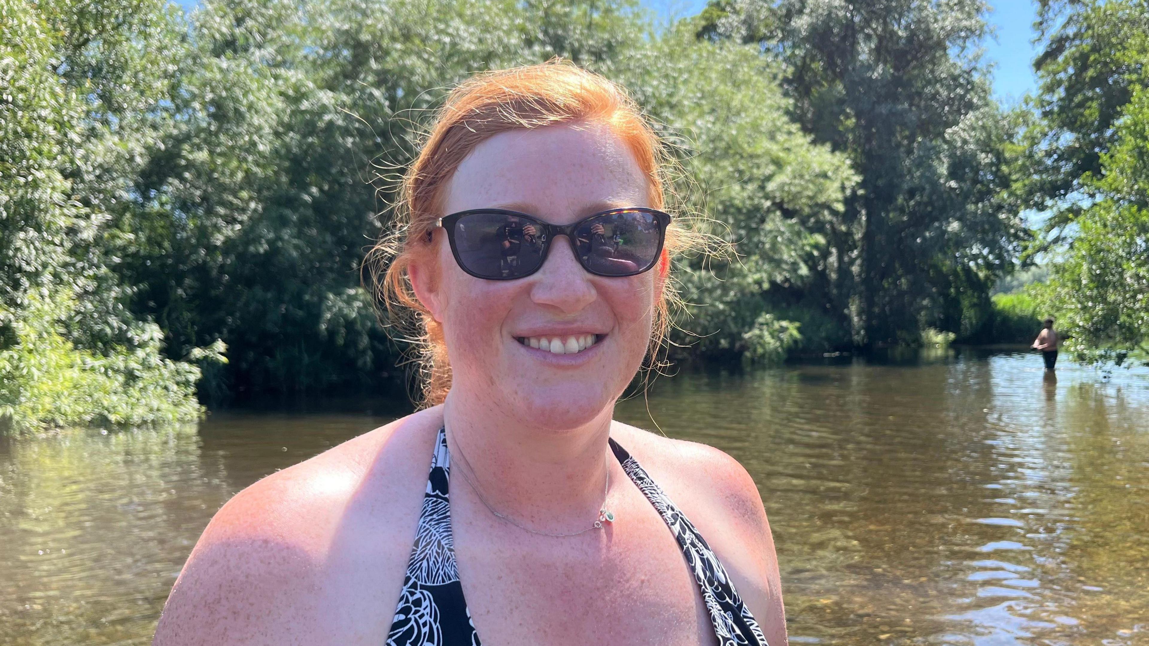 A woman with ginger hair stands in a river wearing sunglasses and a black and white bikini with trees in the background