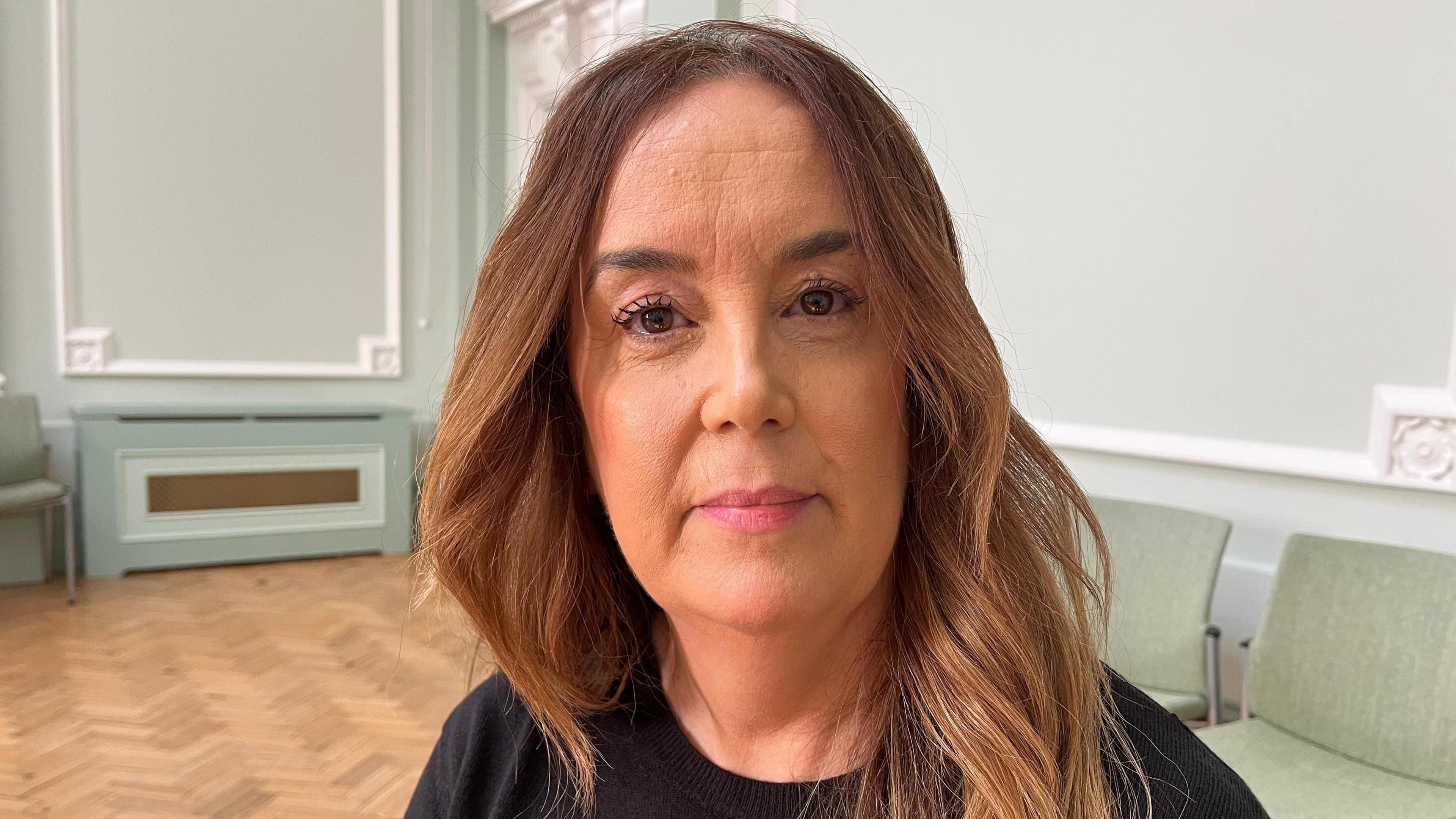 A lady with long brown hair and brown eyes looking into the camera with a slight smile. She is in front of a light green wall, some green chairs and a wooden floor.