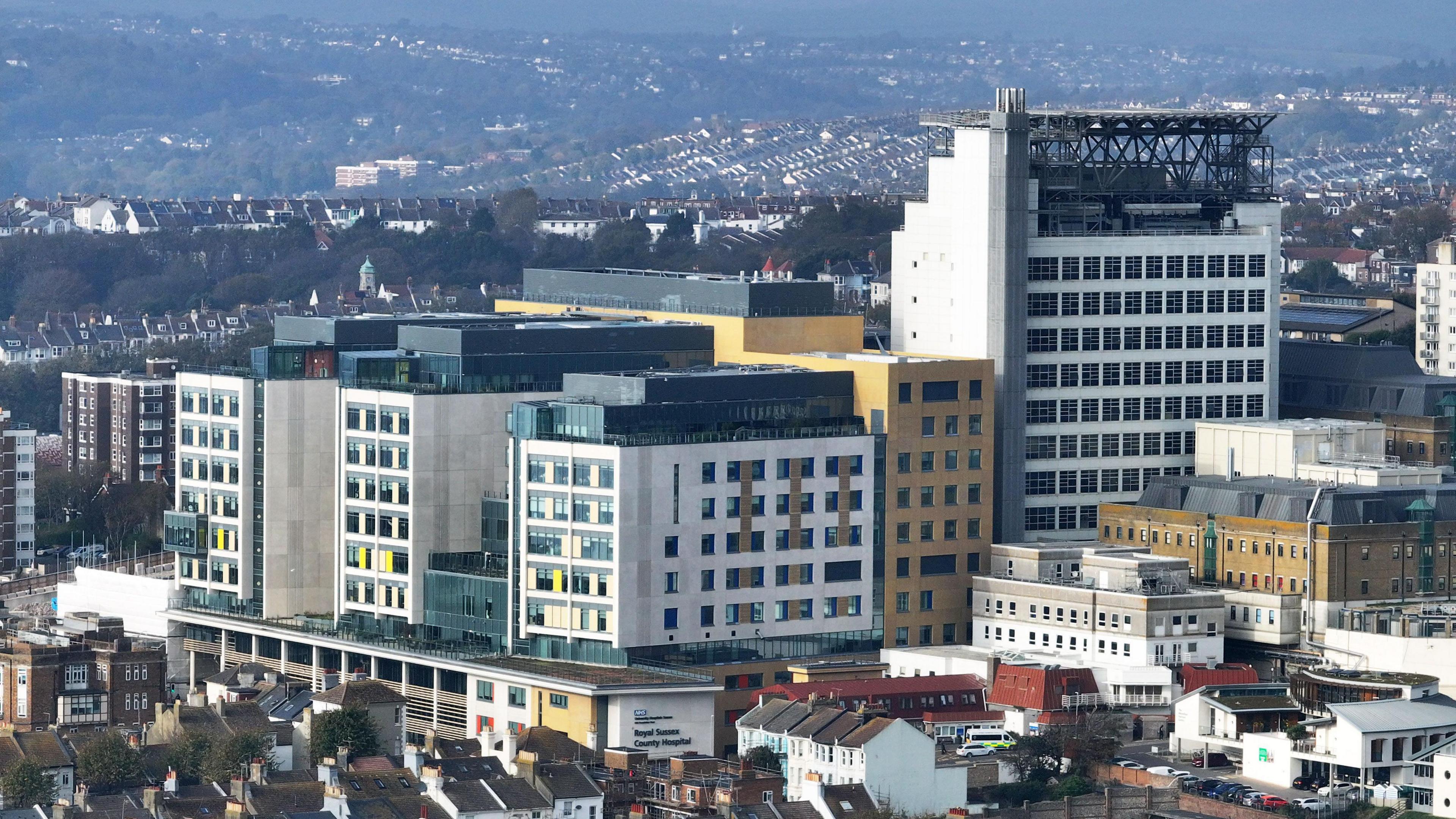 A wide view of Royal Sussex County Hospital. The image shows all the buildings and surrounding houses. The main buildings are white with lots of windows, there is one building in the middle which is bright yellow. 