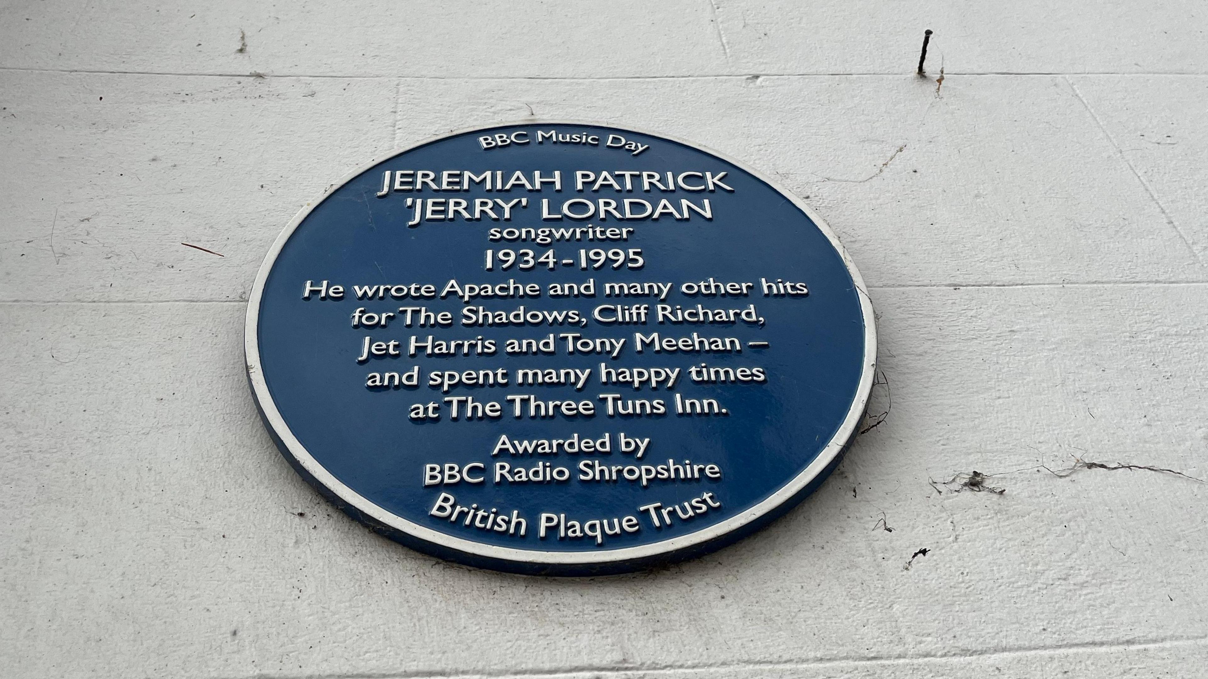 A blue plaque on a whitewashed wall. The plaque has white text reading "BBC Music Day" at the top, and then underneath it reads "Jeremiah Patrick 'Jerry' Lordan', songwriter 1934-1995. He wrote Apache and many other hits for The Shadows, Cliff Richard, Jet Harris, and Tony Meehan - and spent many happy times at The Three Tuns Inn." Beneath that text it reads "Awarded by BBC Radio Shropshire."