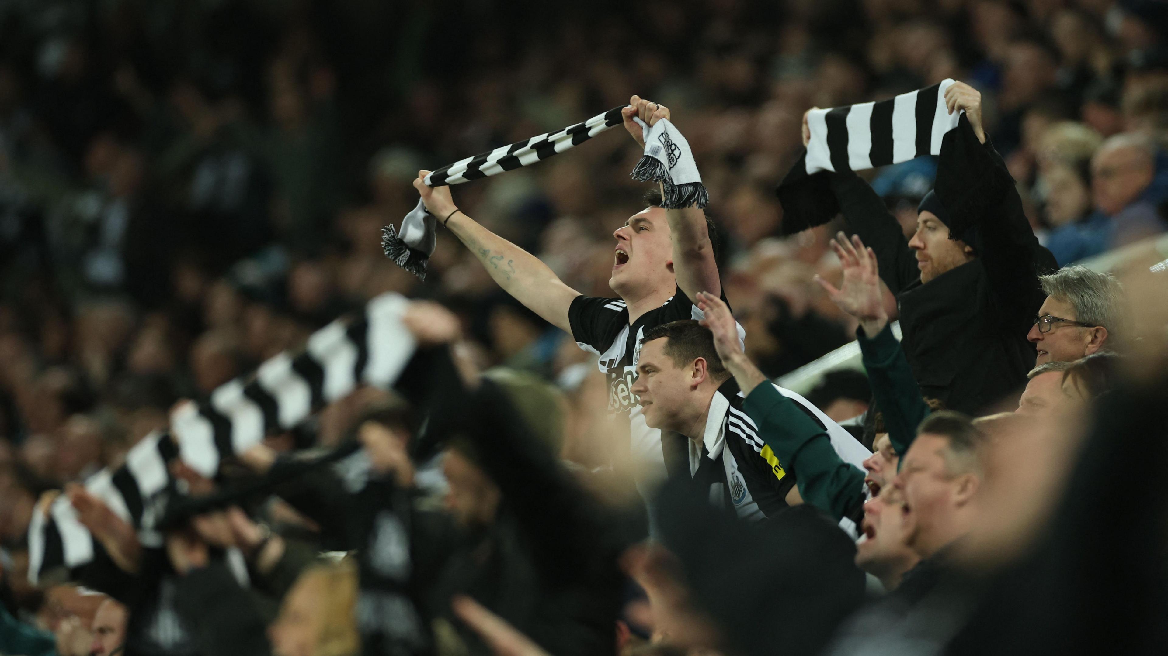 Newcastle United fans wave black-and-white scarves as they cheer their team on during a match at St James' Park. Many are wearing club shirts.