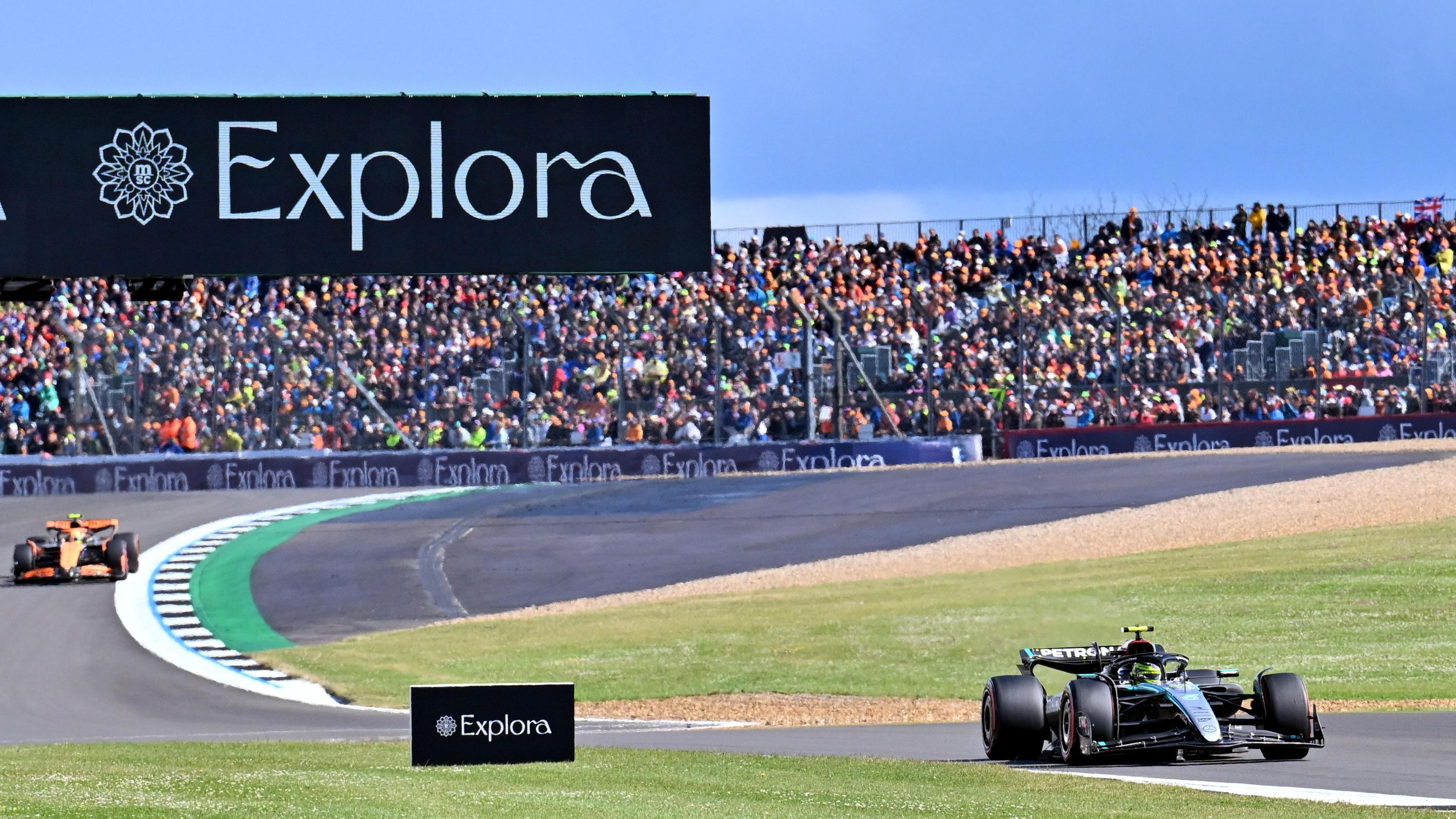 Lewis Hamilton driving on Silverstone Circuit race track with rows and rows of fans dotted behind him. 
