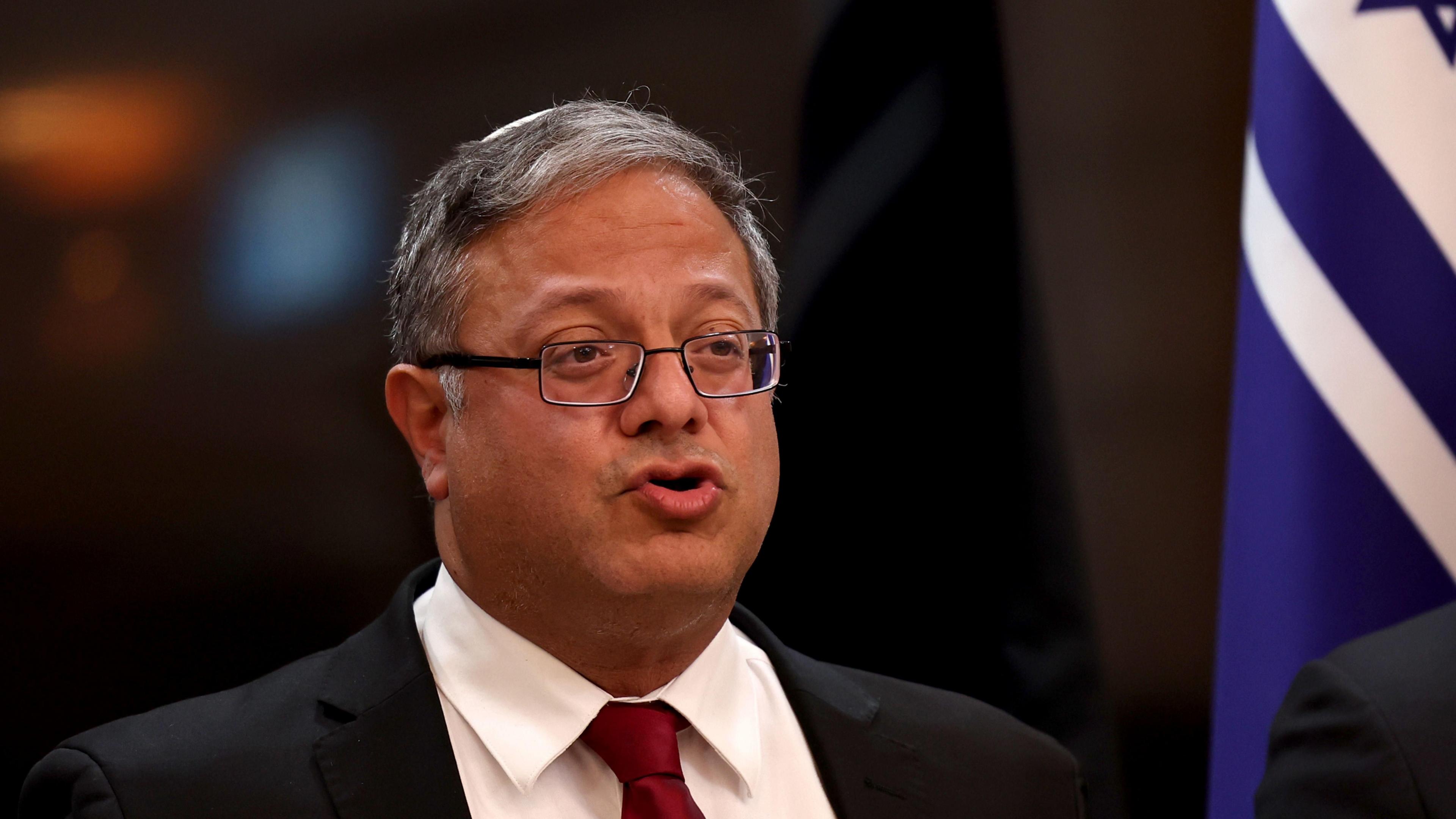 Israeli Minister of National Security Itamar Ben-Gvir delivering a statement to the media, at his ministry headquarters in Jerusalem. He wears glasses, a red tie and white shirt, and stands in front of an Israeli flag.