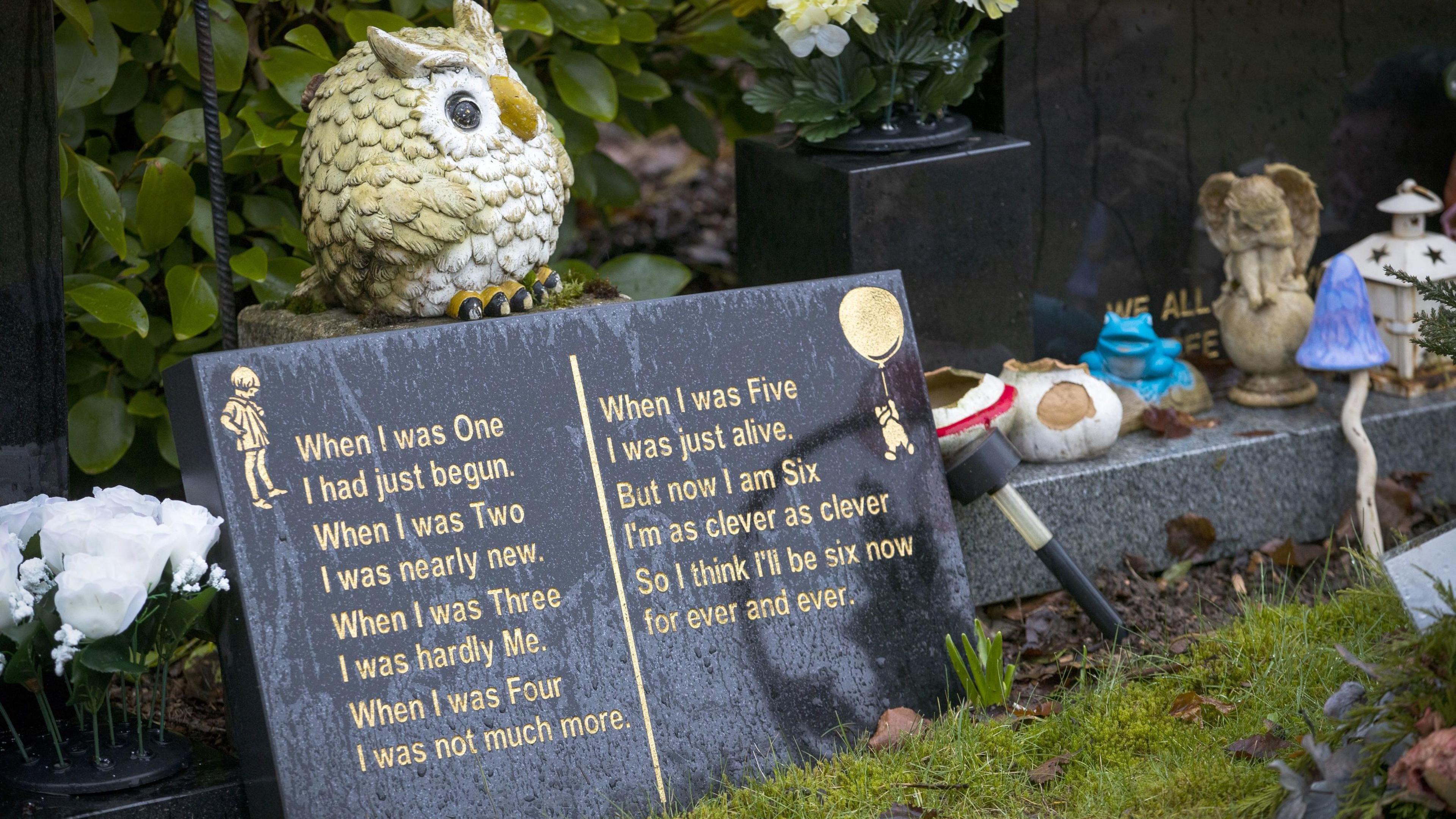 A dark marble memorial plaque with a poem and illustration of a child and Winnie The Pooh. There are toys and little statues scattered around. A statue of an Owl stands on top of the plaque, which sits among grass and trees.