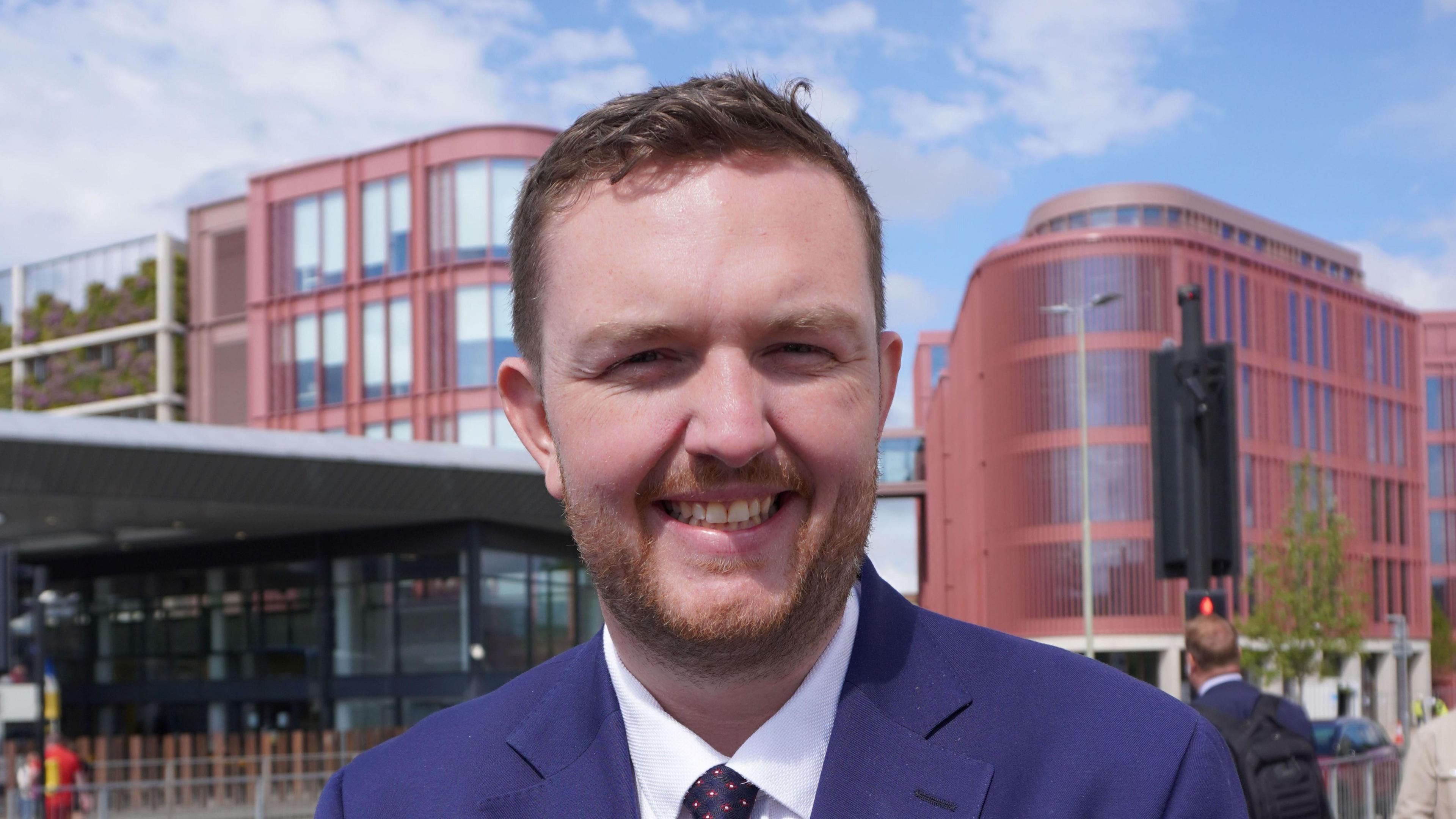 MP Alex McIntyre smiles at the camera while wearing a dark blue suit. He's standing at a traffic junction with a bus station, traffic lights and the new forum development clearly visible in the background.
