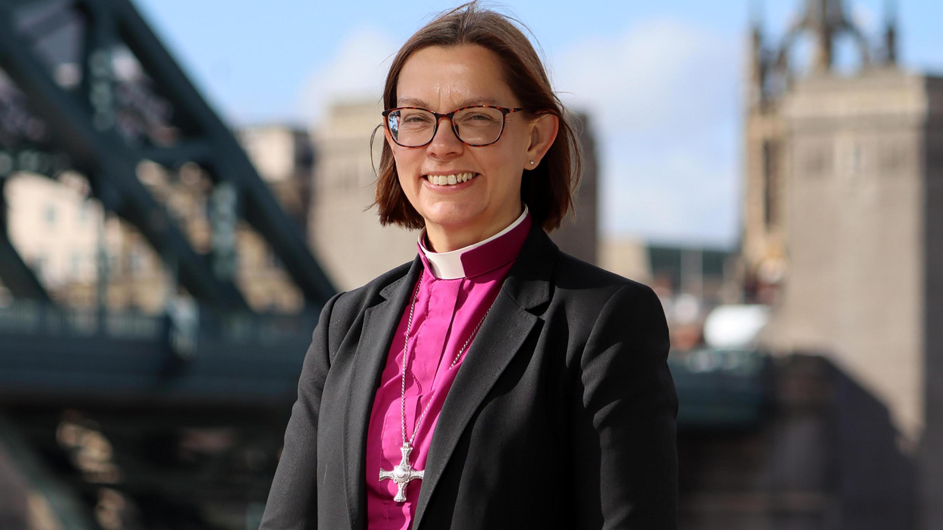 Bishop of Newcastle Helen-Ann Hartley wears a pink robe and brown glasses. Newcastle's Tyne Bridge is in the distance behind her.
