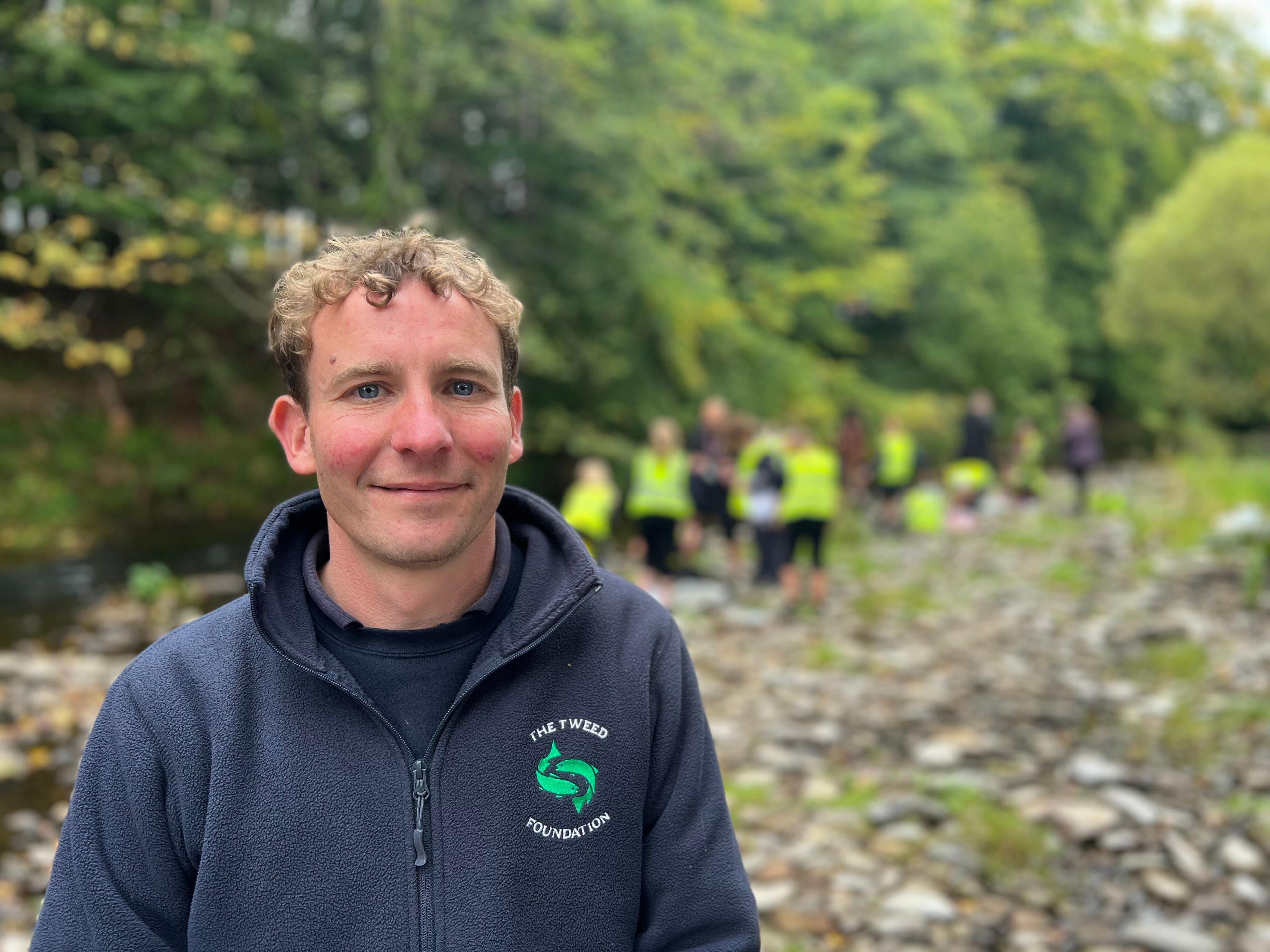 A man with short, blondish, curly hair in a blue zip-top with a green fish logo of the Tweed Foundation. There are children blurred in the background standing on a river bank.