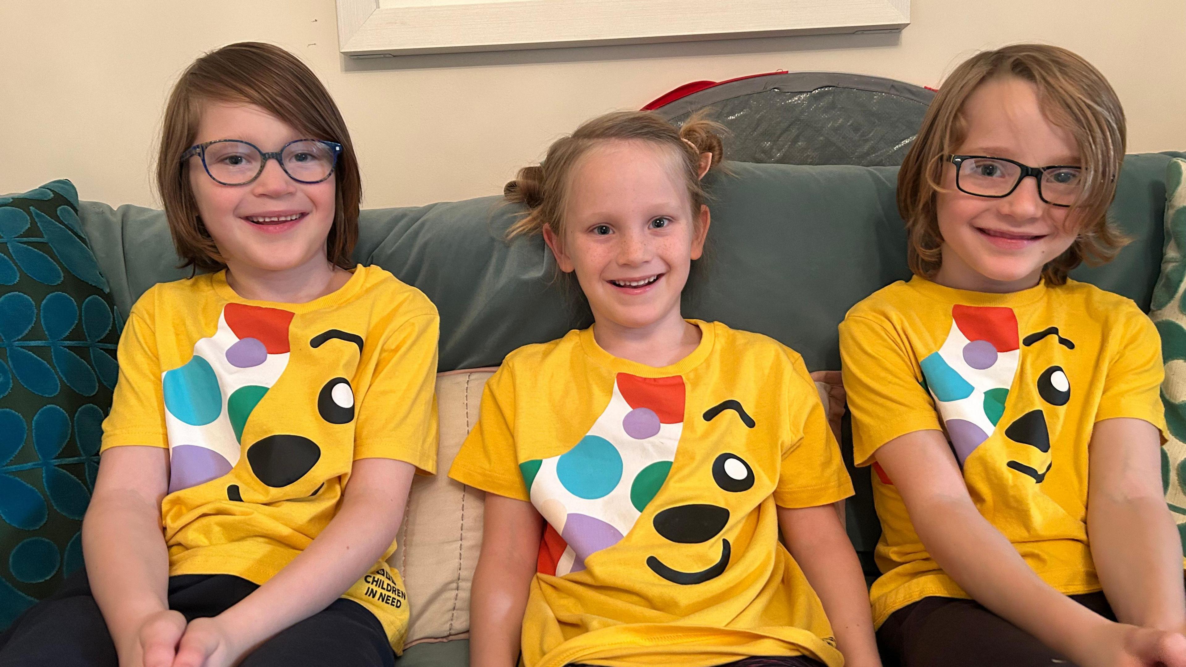 Three children sitting on a coach wearing Pudsey t-shirts and smiling at the camera. two wear glasses.