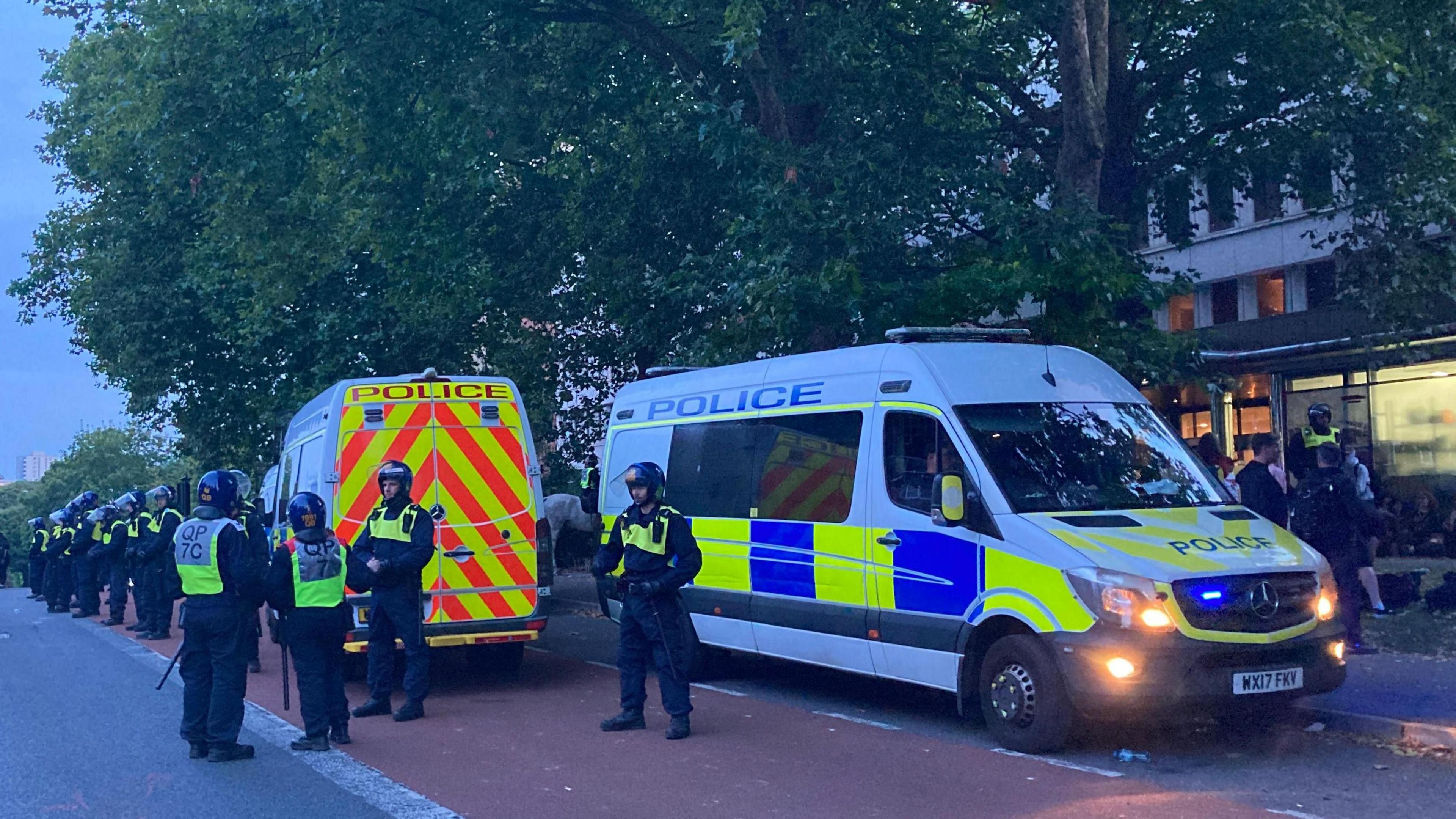 A line of police officers wearing riot gear and helmets outside the Mercure Bristol Hotel. There are several police vans parked outside the entrance, blocking the road. It is dusk and the headlights of the police vans are on.