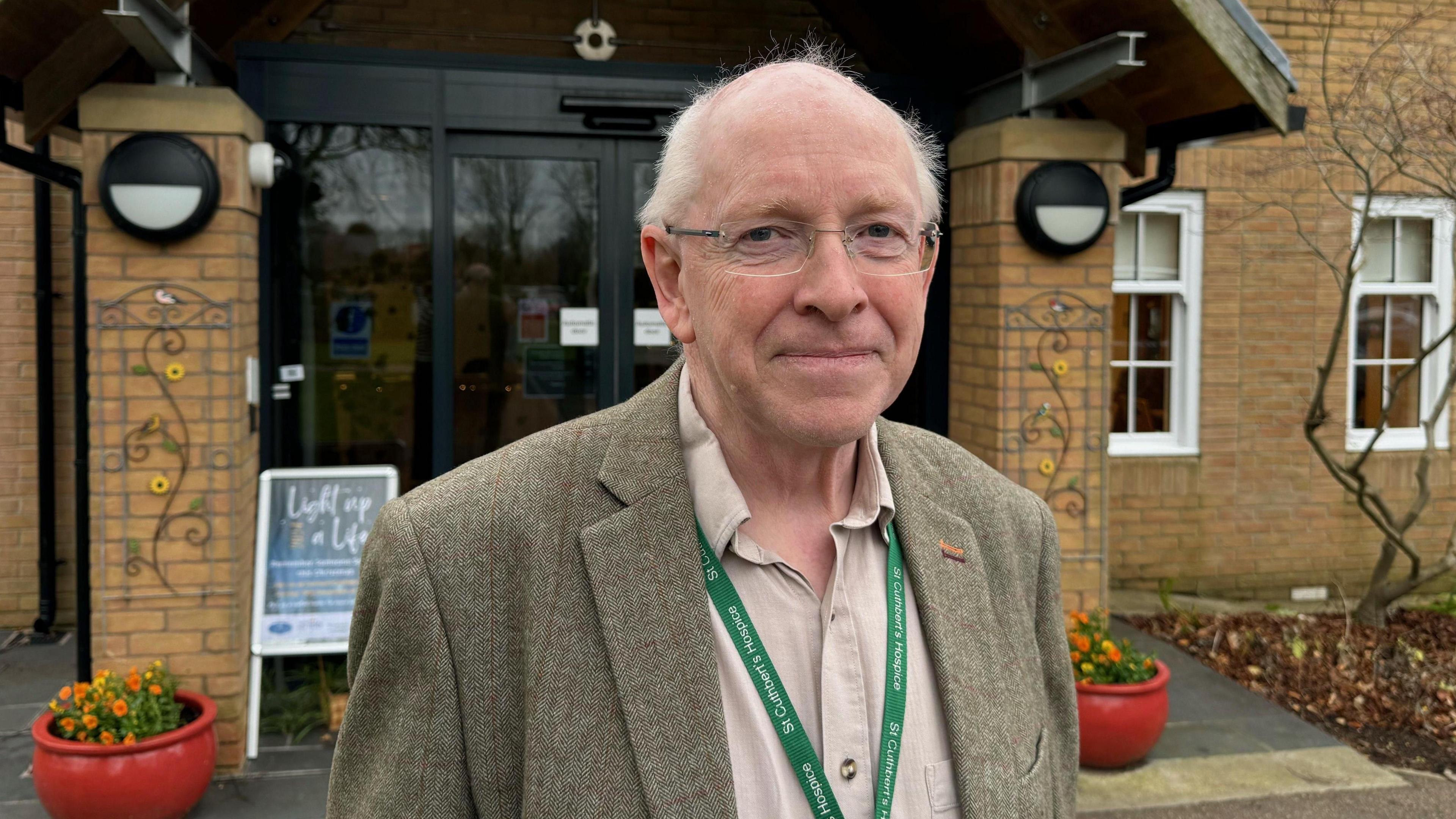 Paul Marriott standing in front of the entrance to St Cuthbert's Hospice. He is bald with short grey hair at the sides and is wearing glasses and a tweed blazer and khaki shirt. The building behind him is made from brick and has glass doors, tinted black.