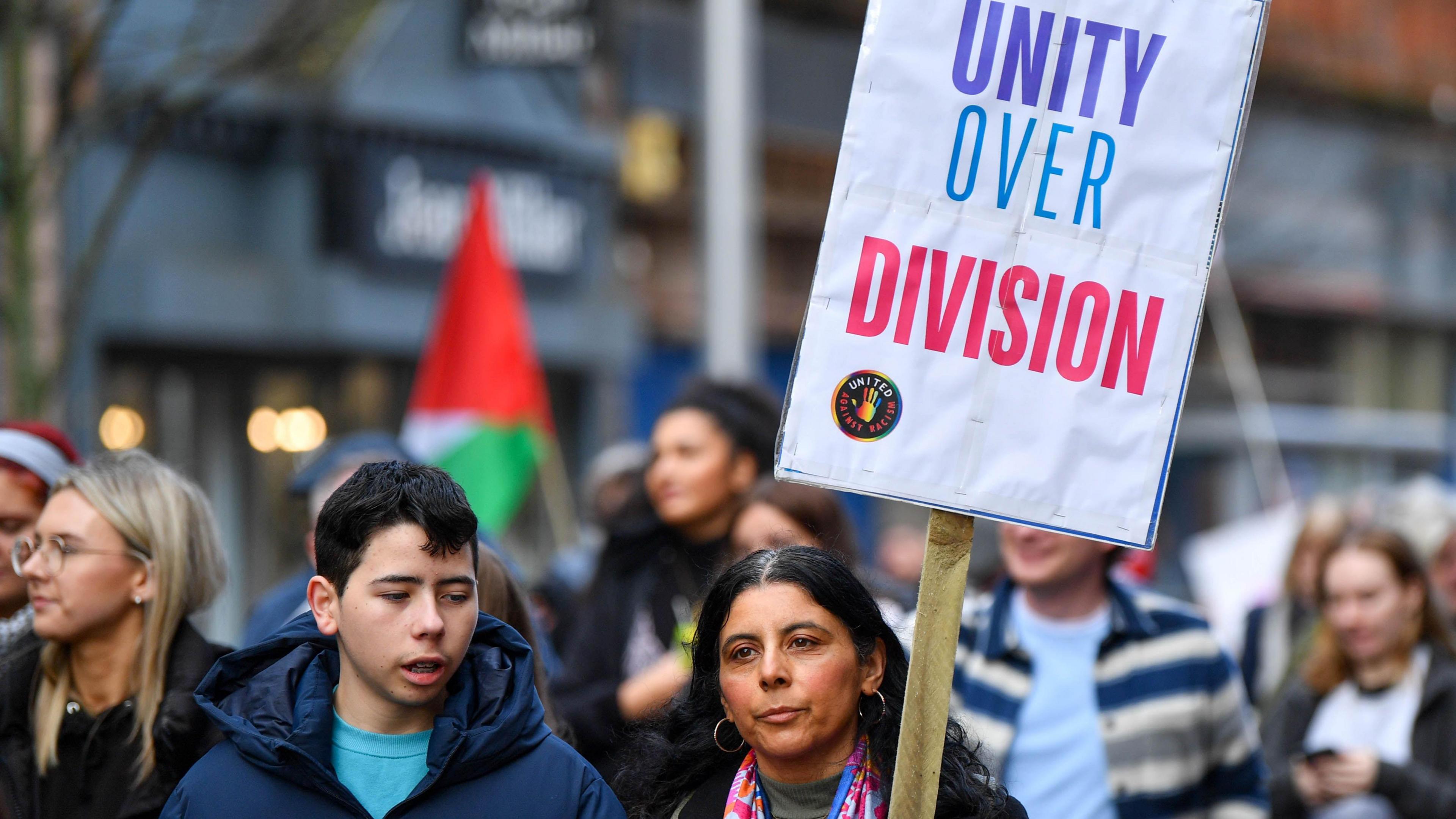 Two protesters who are part of the rally - one is a teenage boy with dark hair and a blue hooded coat, the other is a woman with long dark hair who has a sign which reads: 'Unity over Division'