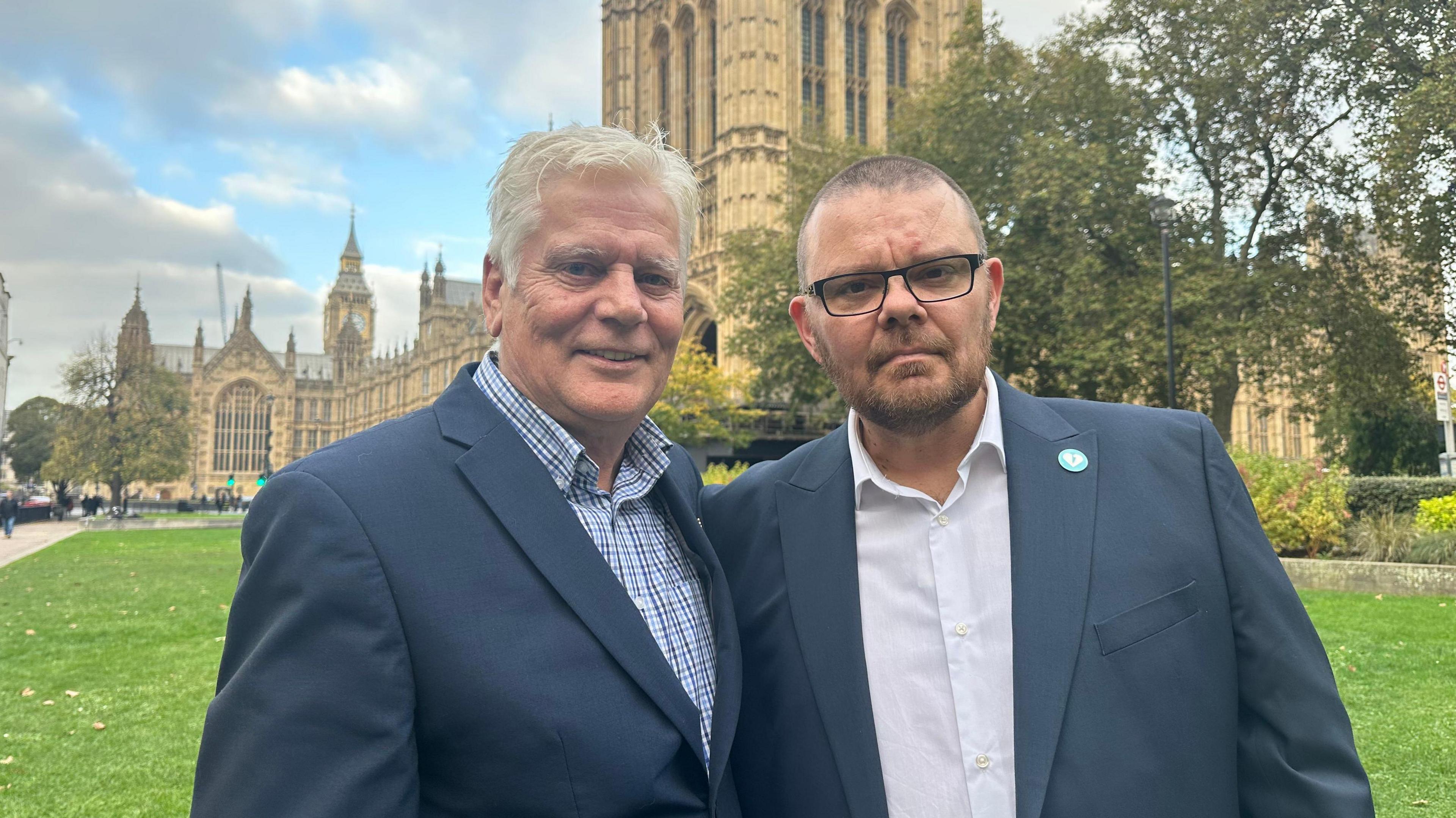 Colin Knox, a white haired man wearing a suit jacket stands next to Martin Cosser, a short haired man with glasses, in front of the Houses of Parliament