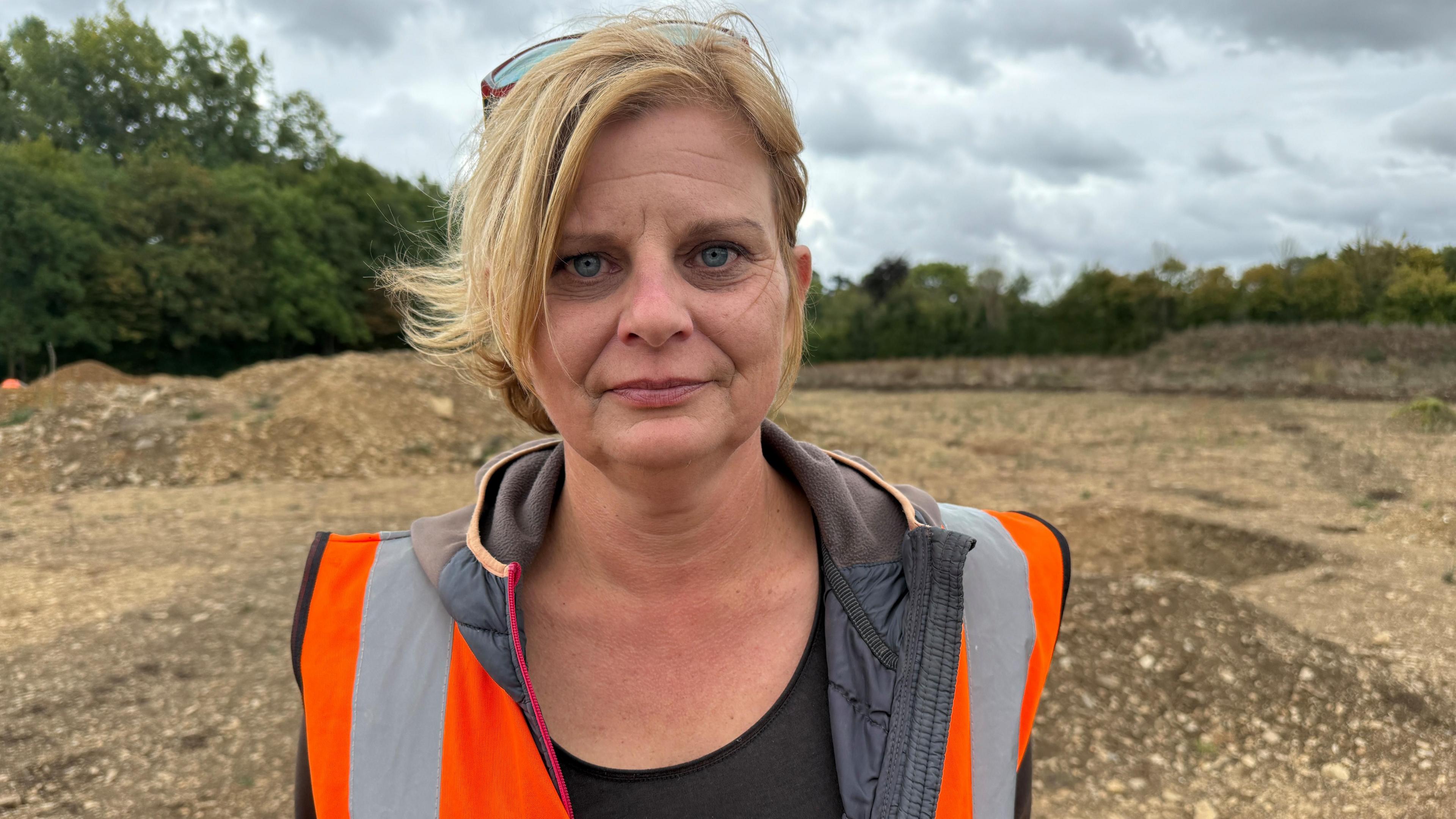 A woman wearing an orange hi-vis vest is standing in a field with the earth dug out behind her. She has blonde hair and is looking at the camera.