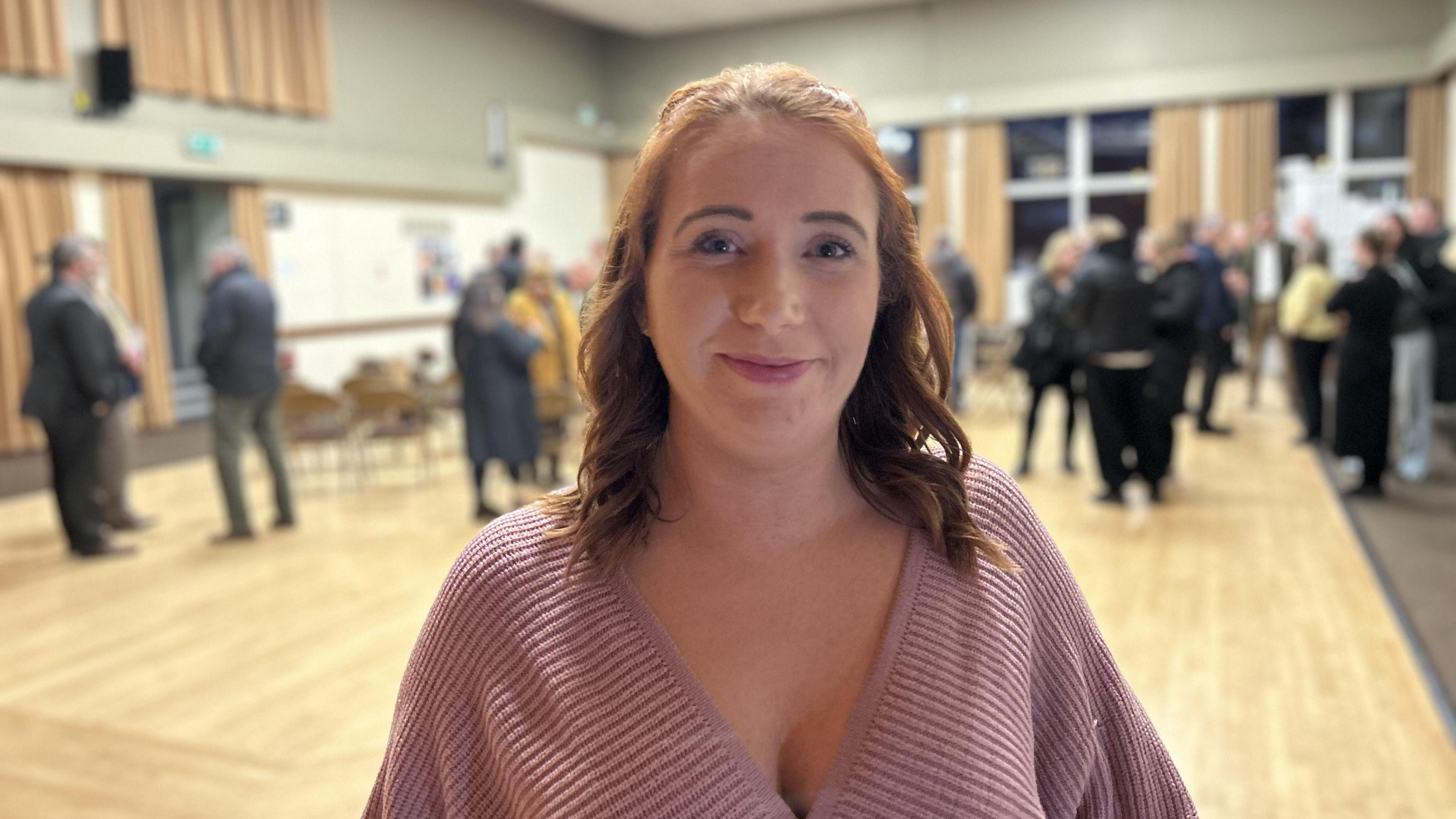 Roseanne Plater smiles at the camera at a public meeting. She has red-brown hair and is wearing a pink top. A crowd of people are in the hall behind her.