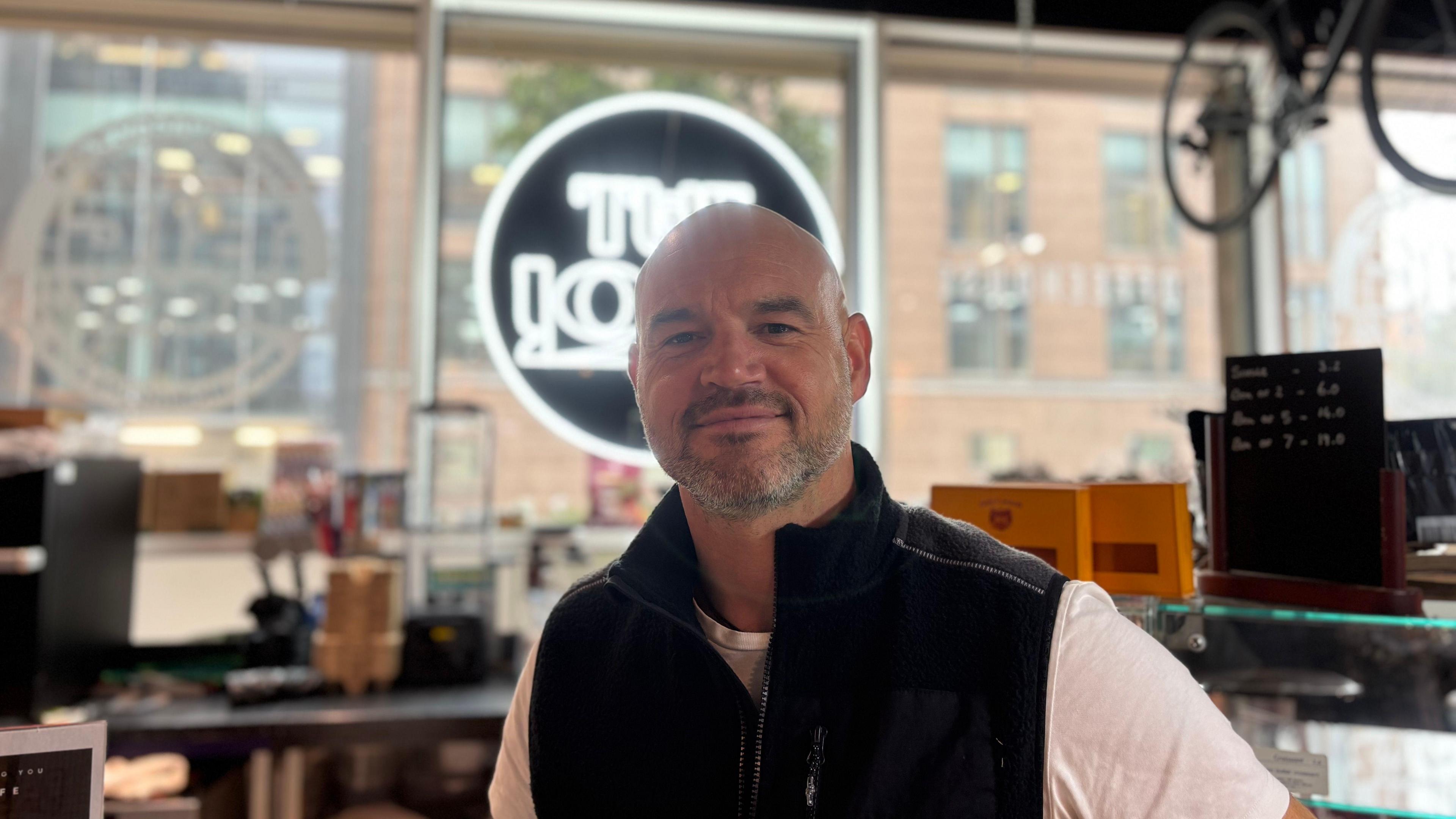 A picture of Dave stood in front of a camera smiling at it. He is wearing a white shirt with a black gilet and he has a beard. Behind him is also a bar with his company logo shining behind his head.