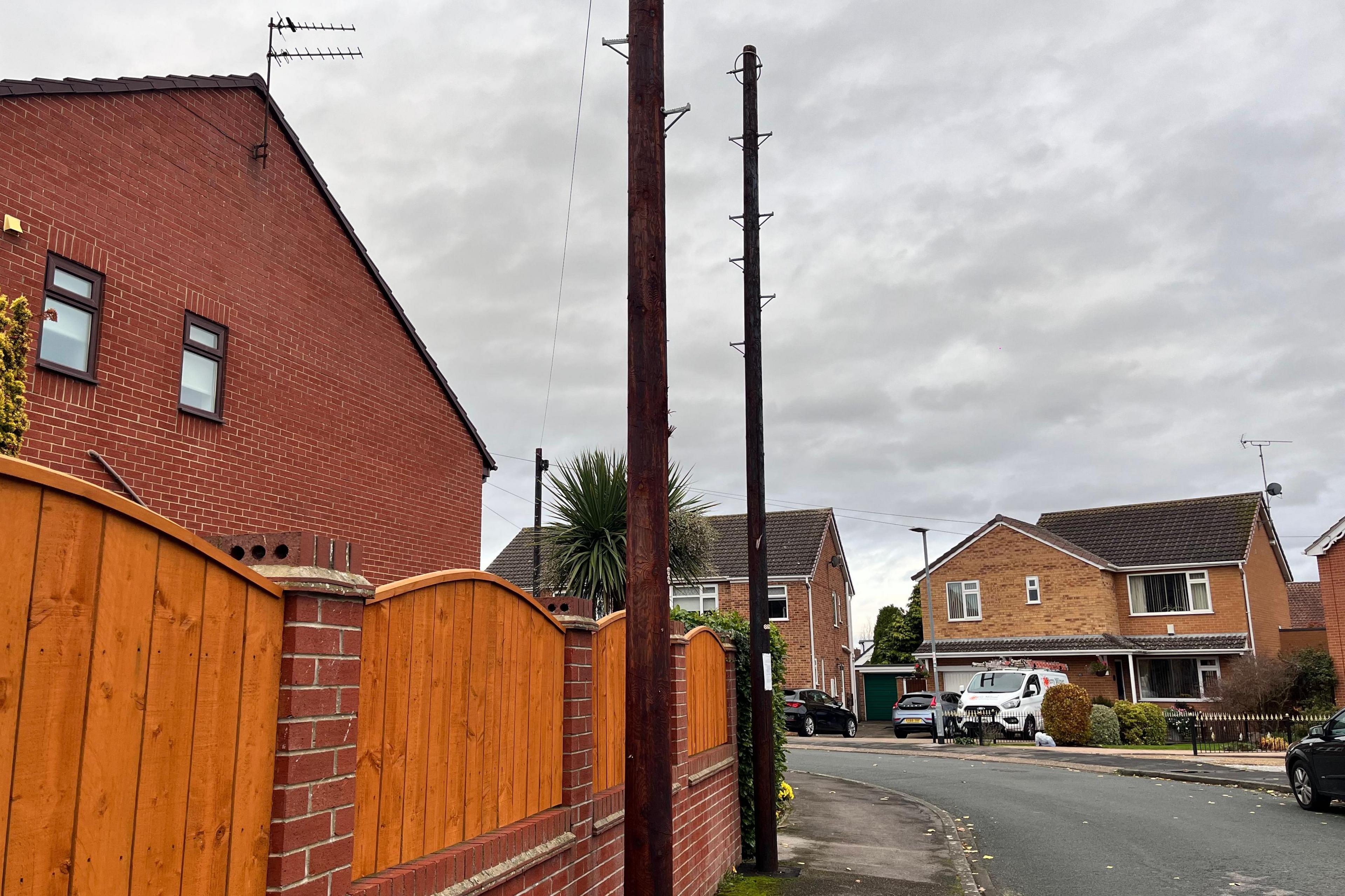 Two new brown broadband poles stand tall on a pavement outside homes in a residential area in Cottingham. They are installed close to a light-brown fence, close to the garden fence of a home. One is wired up, the other is not. A street sweeps round in front of other houses.