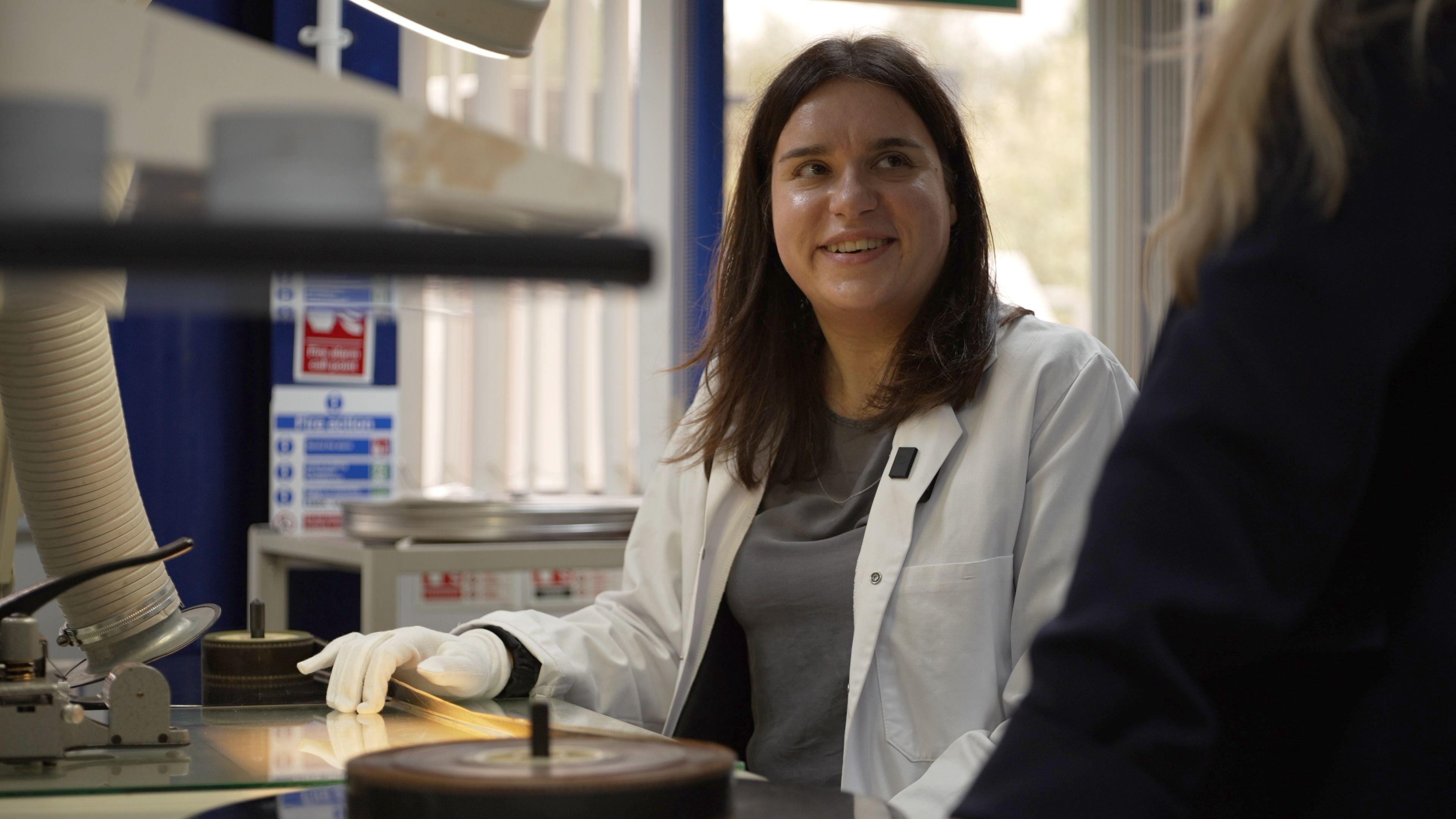 Elena Nepoti, a woman wearing white gloves and lab coat, sits at a desk with a light shining up from it. A reel of film is in the foreground and stretched to another reel 