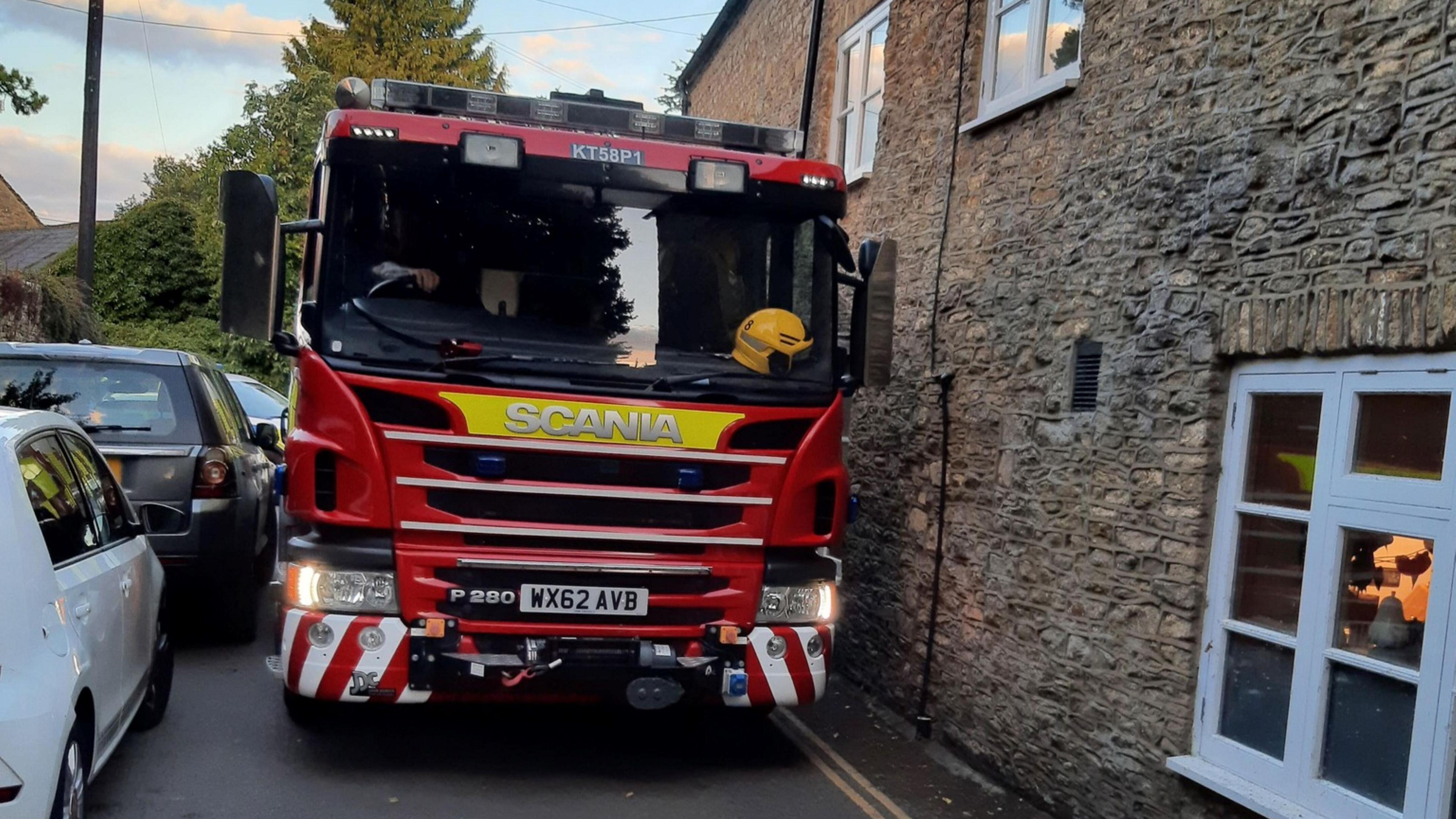 A fire engine squeezes between a stone wall and a series of cars parked on the other side of the road. Trees and a pole in the background.