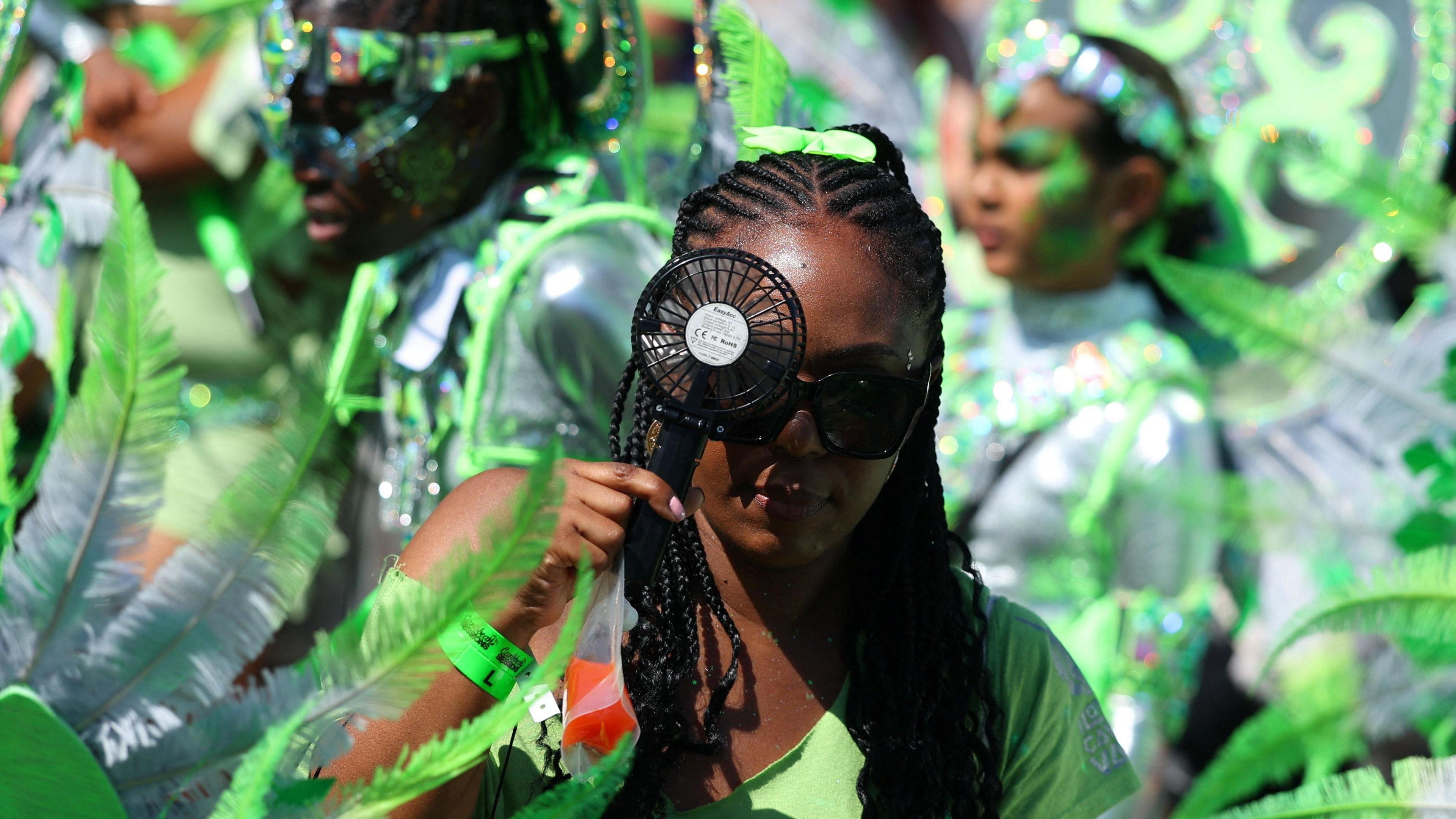 A festival-goer in a sea of green-clad performers looks at the camera and holds a battery operated fan close to her face.