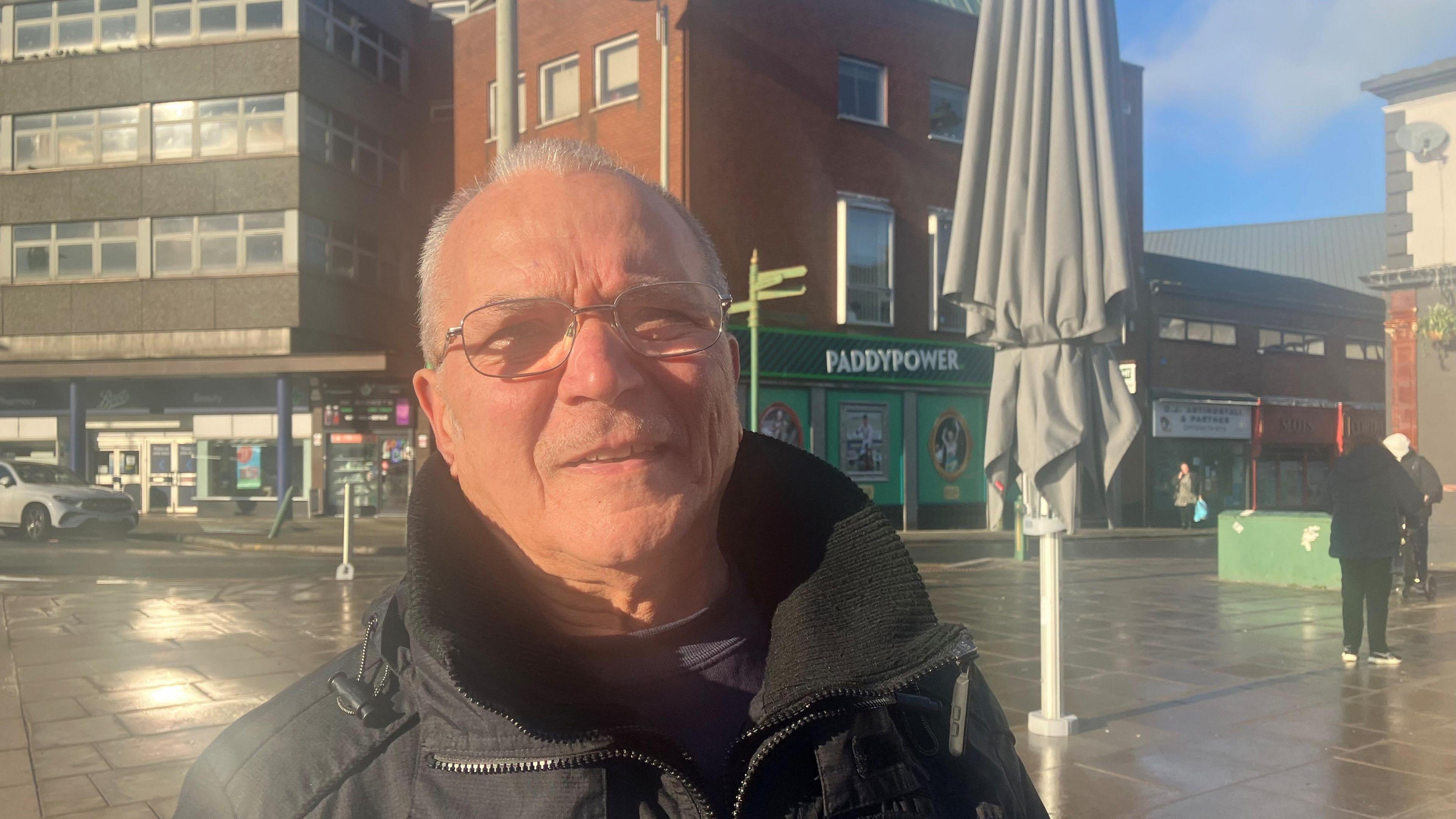 Alfredo Pisani, who is wearing a black coat and has glasses, stands in front of one of the grey parasols. Shops and traffic can be see in the distance with people walking around the area. 