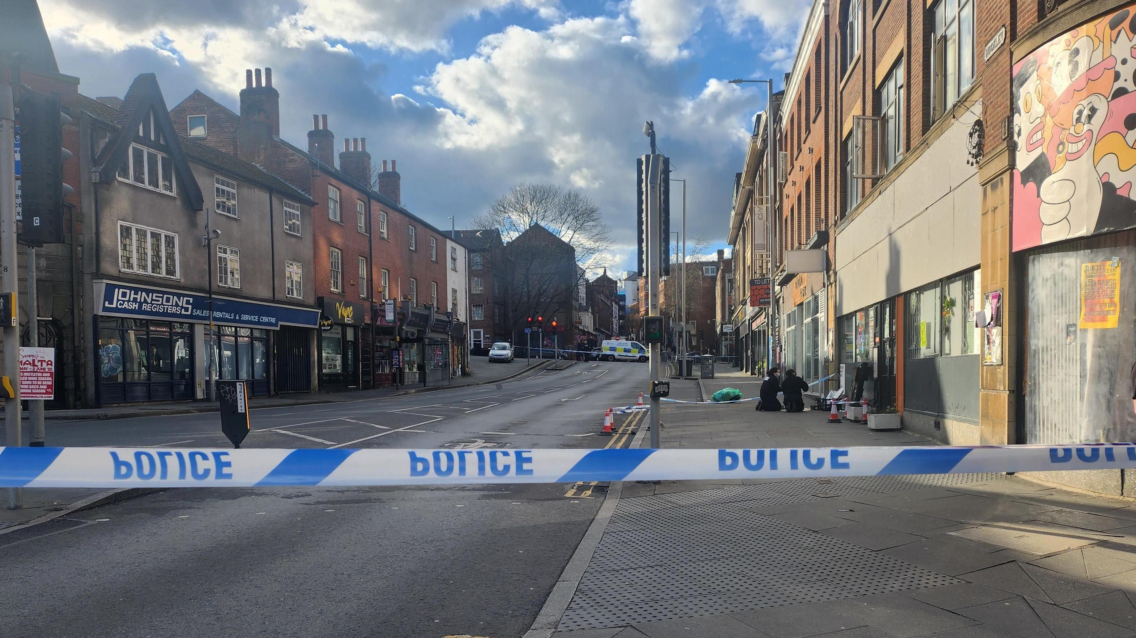 Blue and white "Police" tape is across both ends of the road. A police officer is stood just behind the tape to the right of the picture. Two other officers are kneeling on the floor in the background.