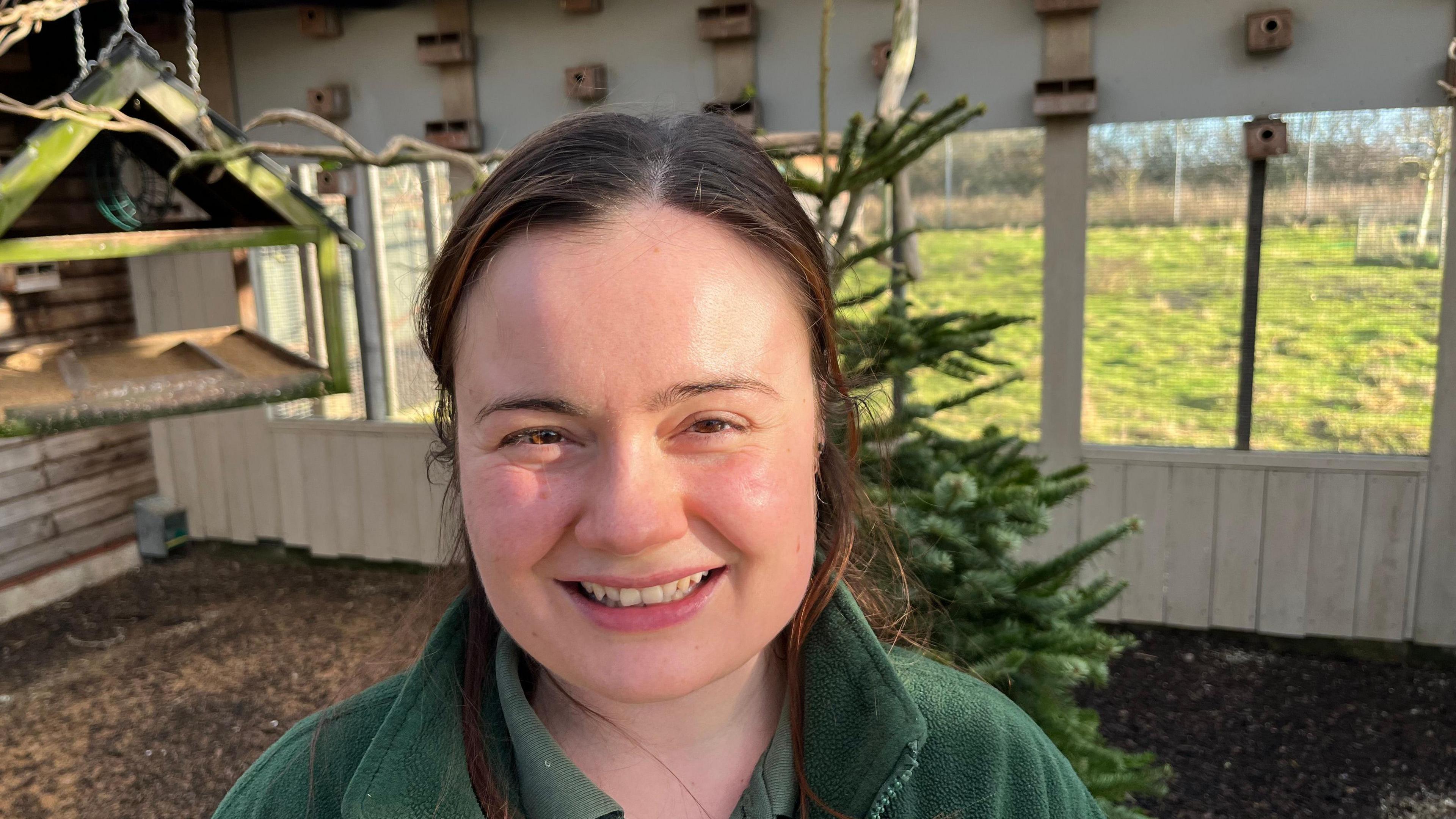 Cathy Evans smiles at the camera. She has dark hair and is wearing a green fleece with a matching shirt beneath. She is standing in a bird aviary with a Christmas tree visible behind her.