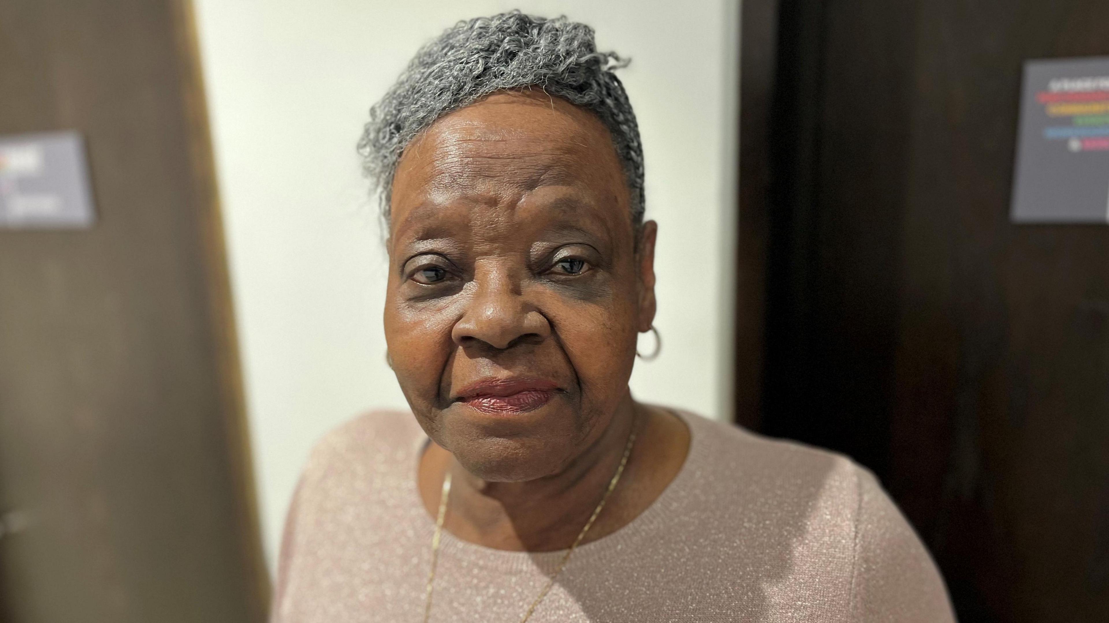 Vilma stands in front of a cream-coloured wall with brown doors to the left and right in the background. She is wearing a beige round-neck top with a long thin gold chain around her neck and small gold hoop earrings. She has greying hair pulled back from her face.