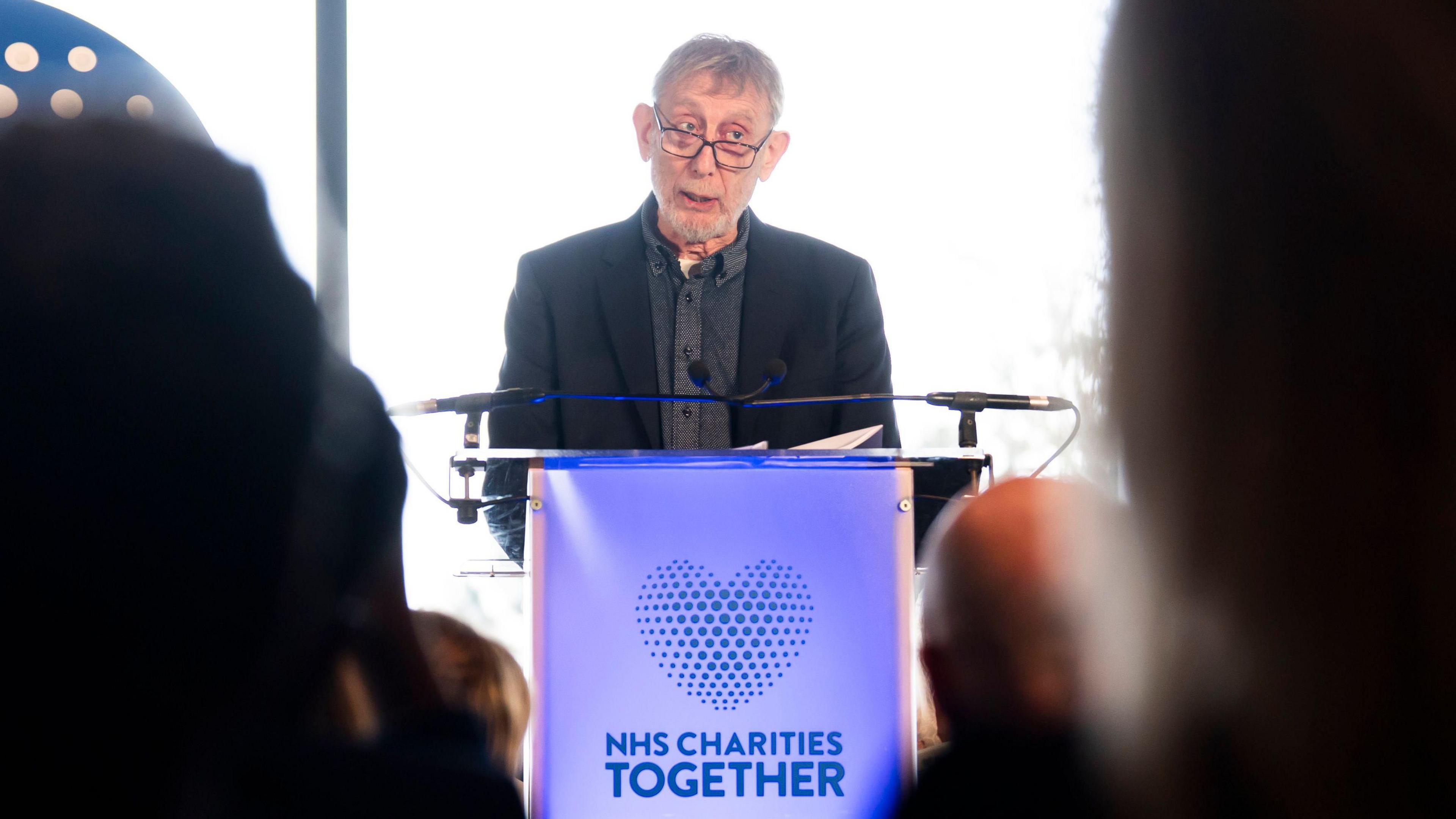 Michael Rosen, a man wearing a black suit and grey shirt, is stood at a blue lectern which has the words NHS Charities Together written on it along with a heart symbol.