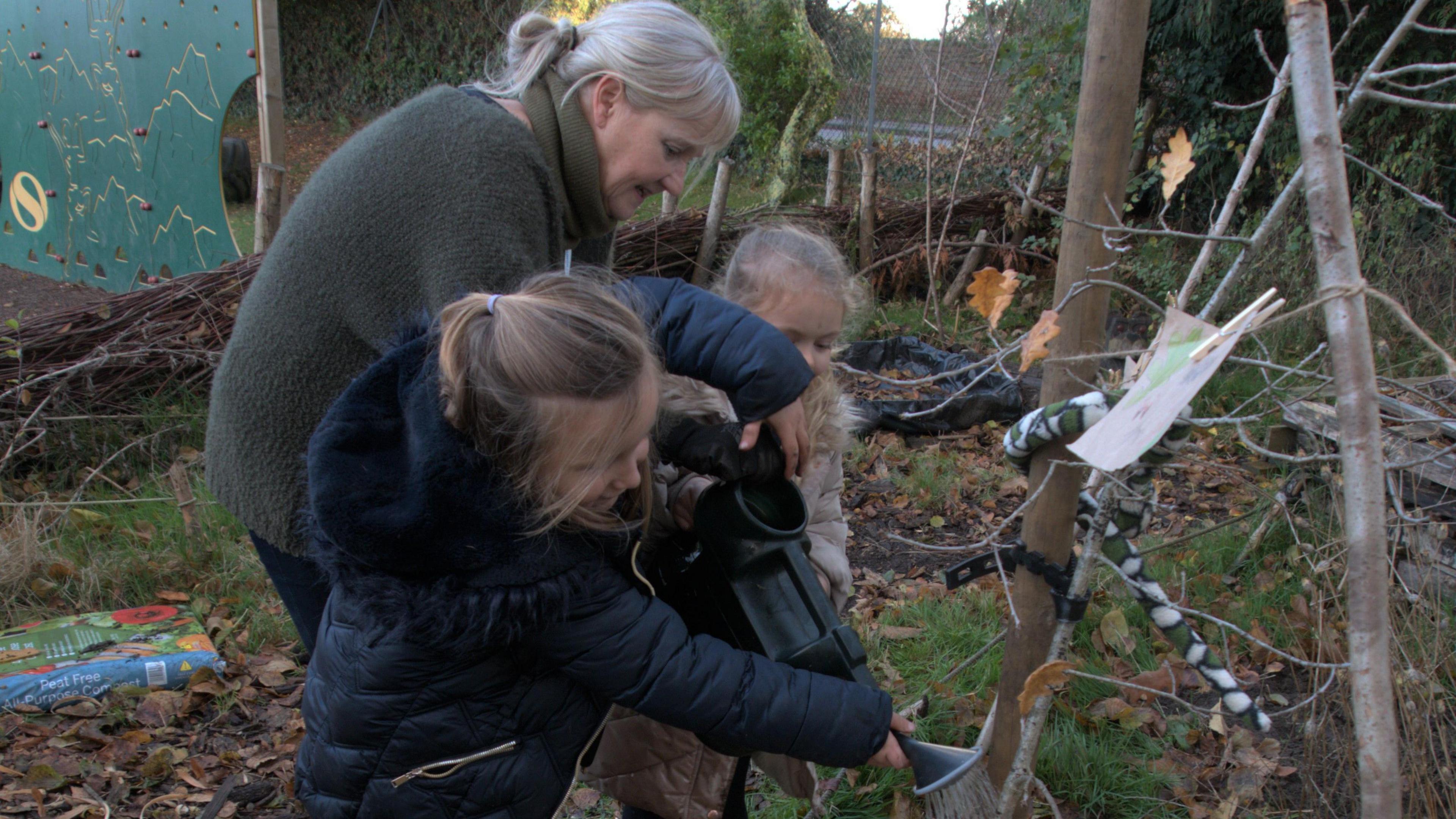 In a rural setting, Green councillor Sally Noble, wearing a dark green jumper, is helping two pupils with long blond hair water a young oak tree with a watering can