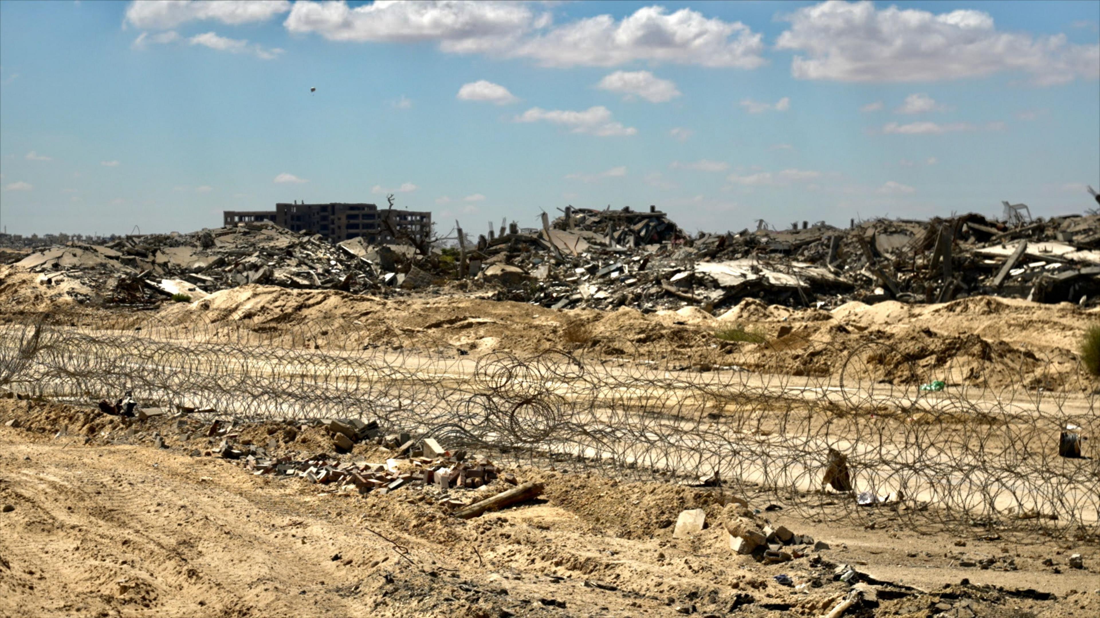 A coil of barbed wire runs across churned-up piles of soil near the city of Rafah. Demolished buildings lie in piles of debris behind the wire, with one lone structure still upright but appearing to have had its windows blown out.  