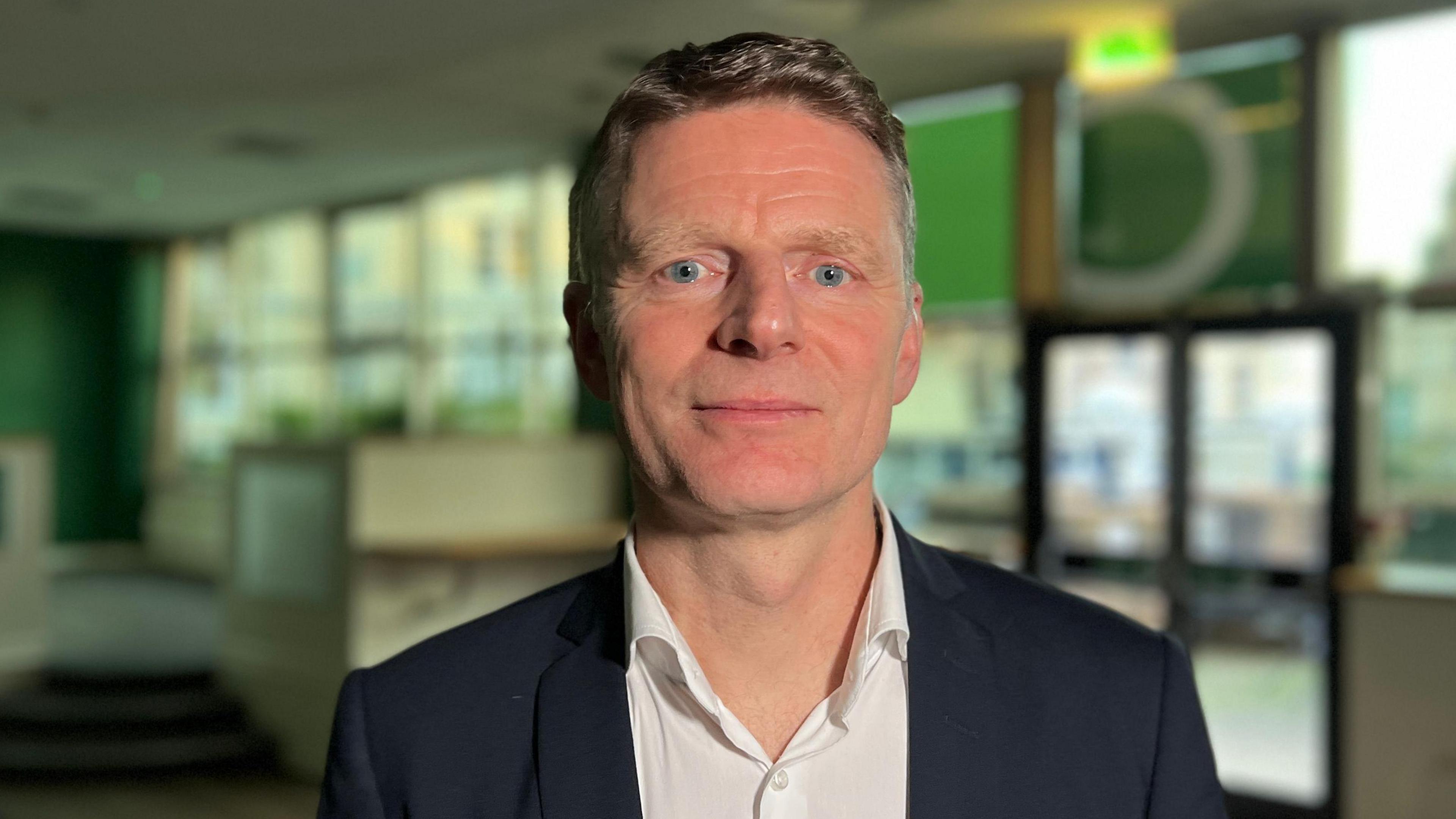 James Hewes, a man with short blond hair, looks into the camera. He is wearing a black blazer and an open white shirt.
