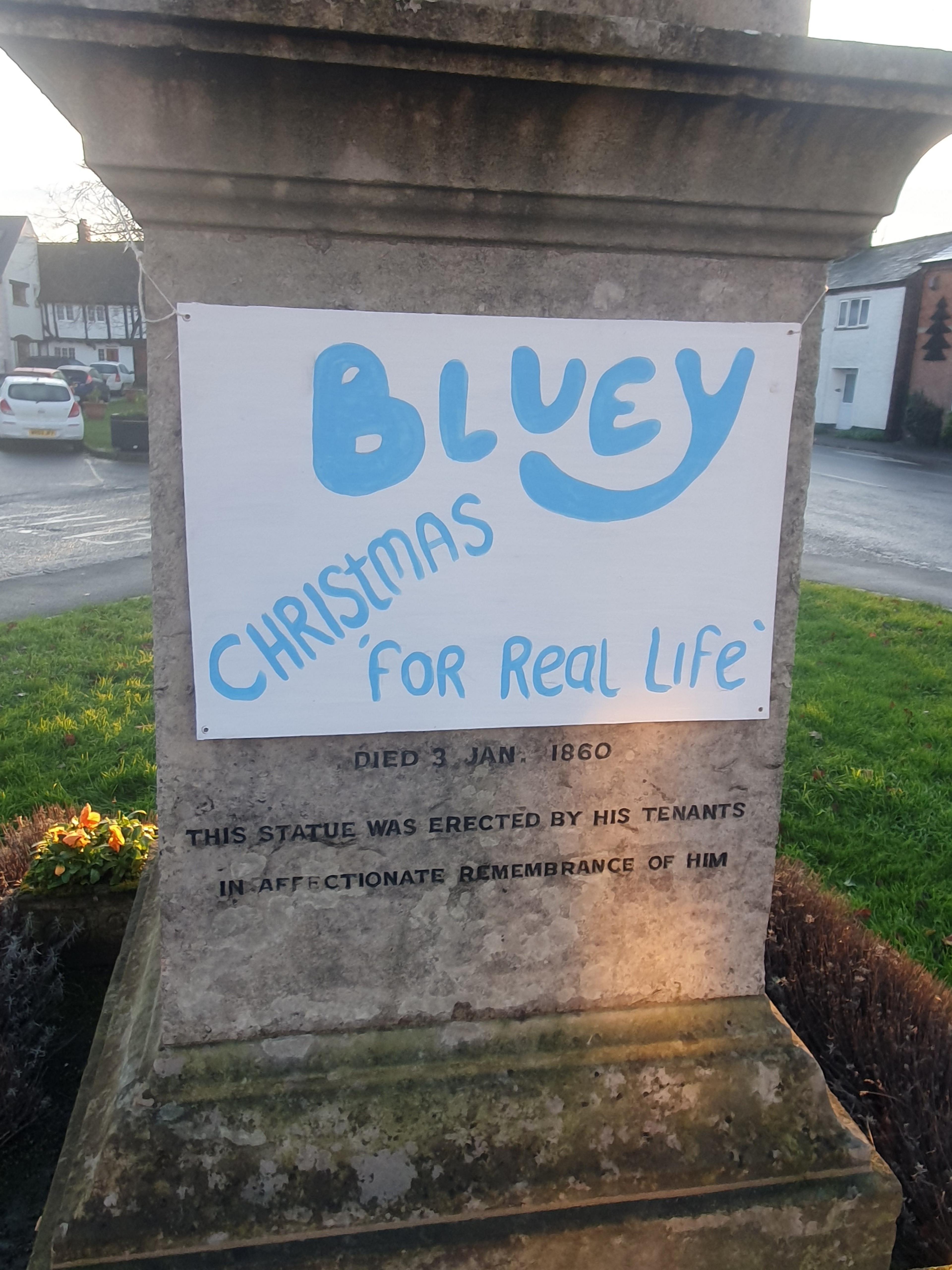 A white placard tied to the statue's plinth, with blue text reading: "Bluey - Christmas - for real life"