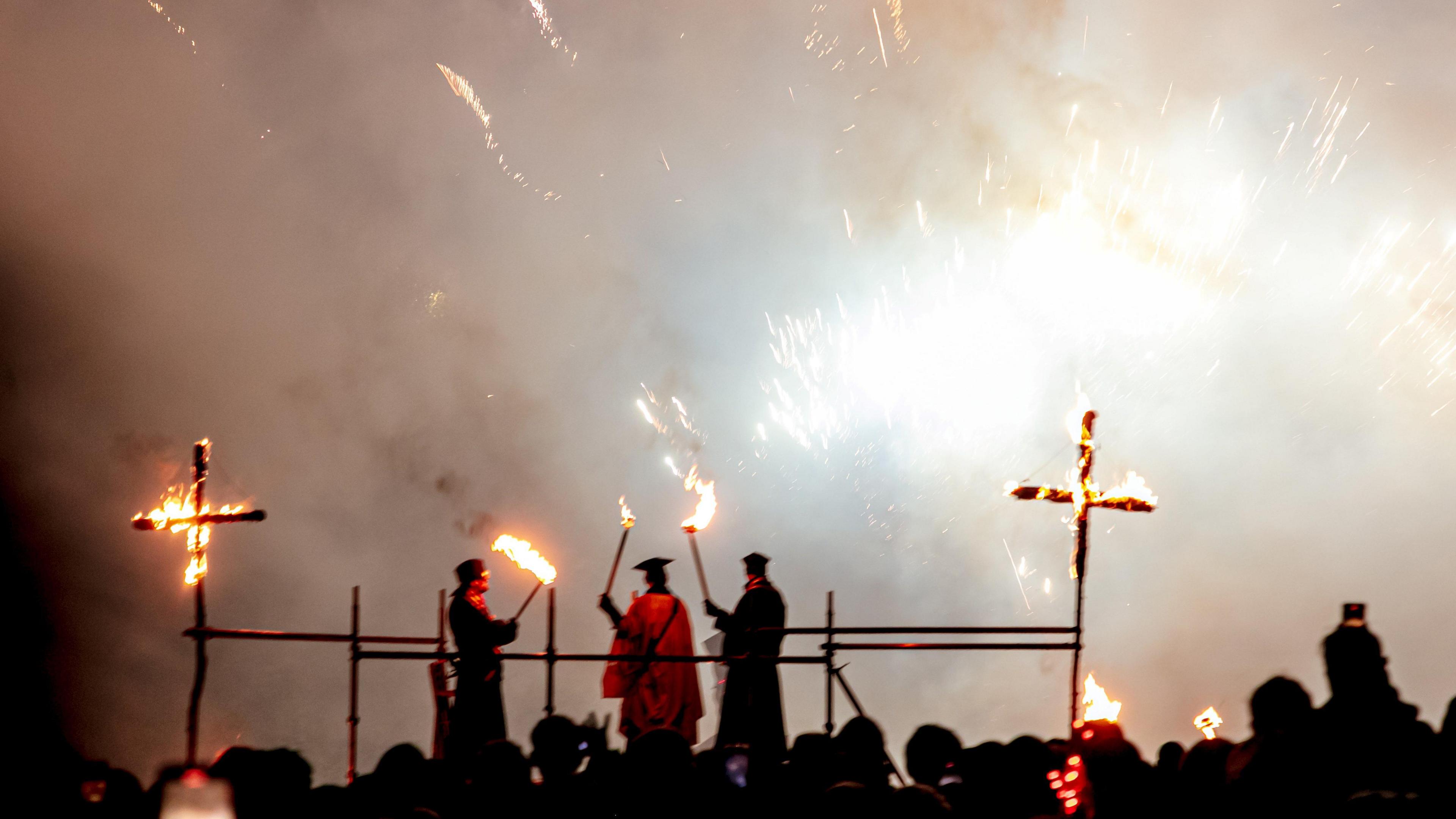 Crosses on fire. The crosses are made out of wood and being burnt by three individuals. There is a large crowd of people watching. 