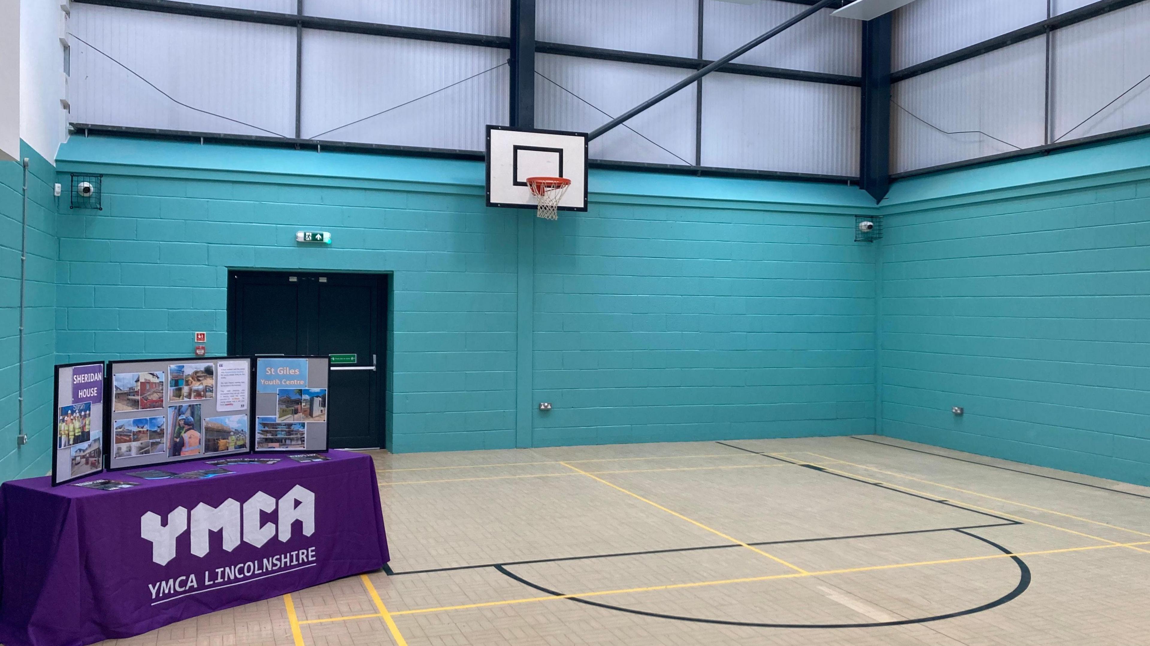 A sports hall with a basketball hoop and pale blue walls. There is a table covered with a purple cloth. Written on the cloth is "YMCA Lincolnshire", and on the table is a display with photographs