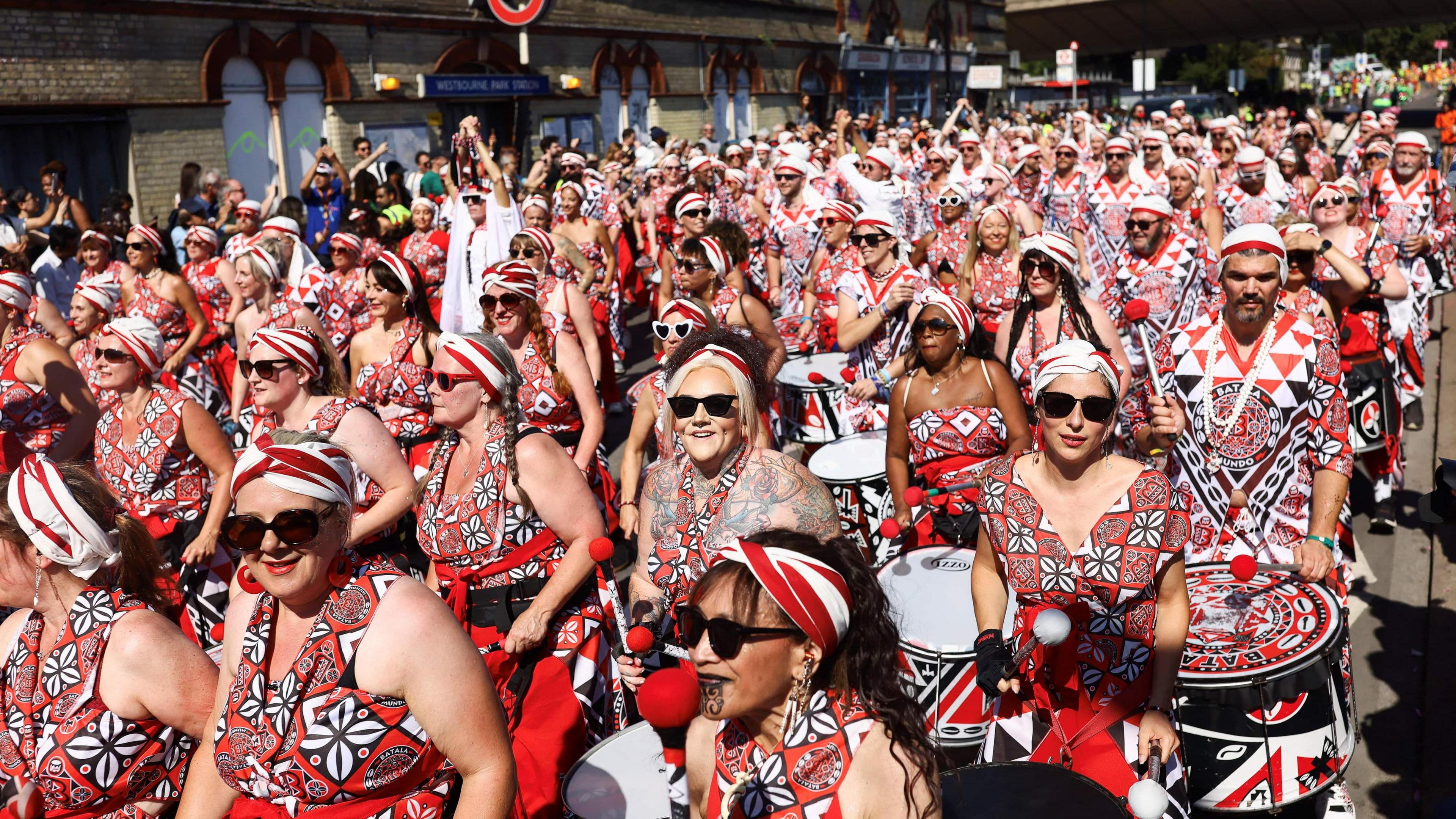 A group of performers in red, black and white patterned clothing, banging drums during the parade.
