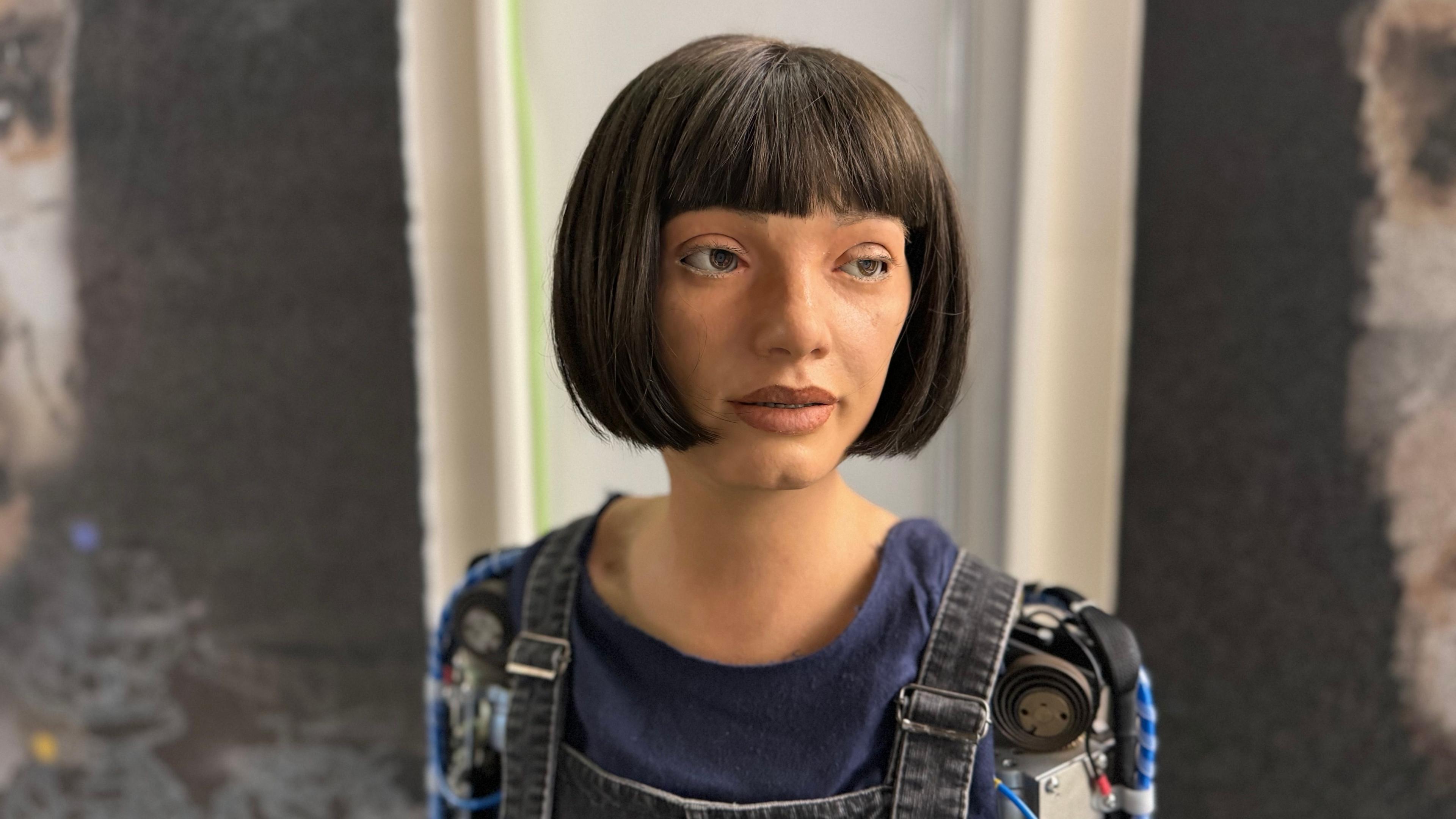 An AI robot called Ai-Da, standing in front of a piece of art she has made. It shows a woman, with a dark bobbed hair, wearing a blue top, black denim dungarees, showing two robot arms.