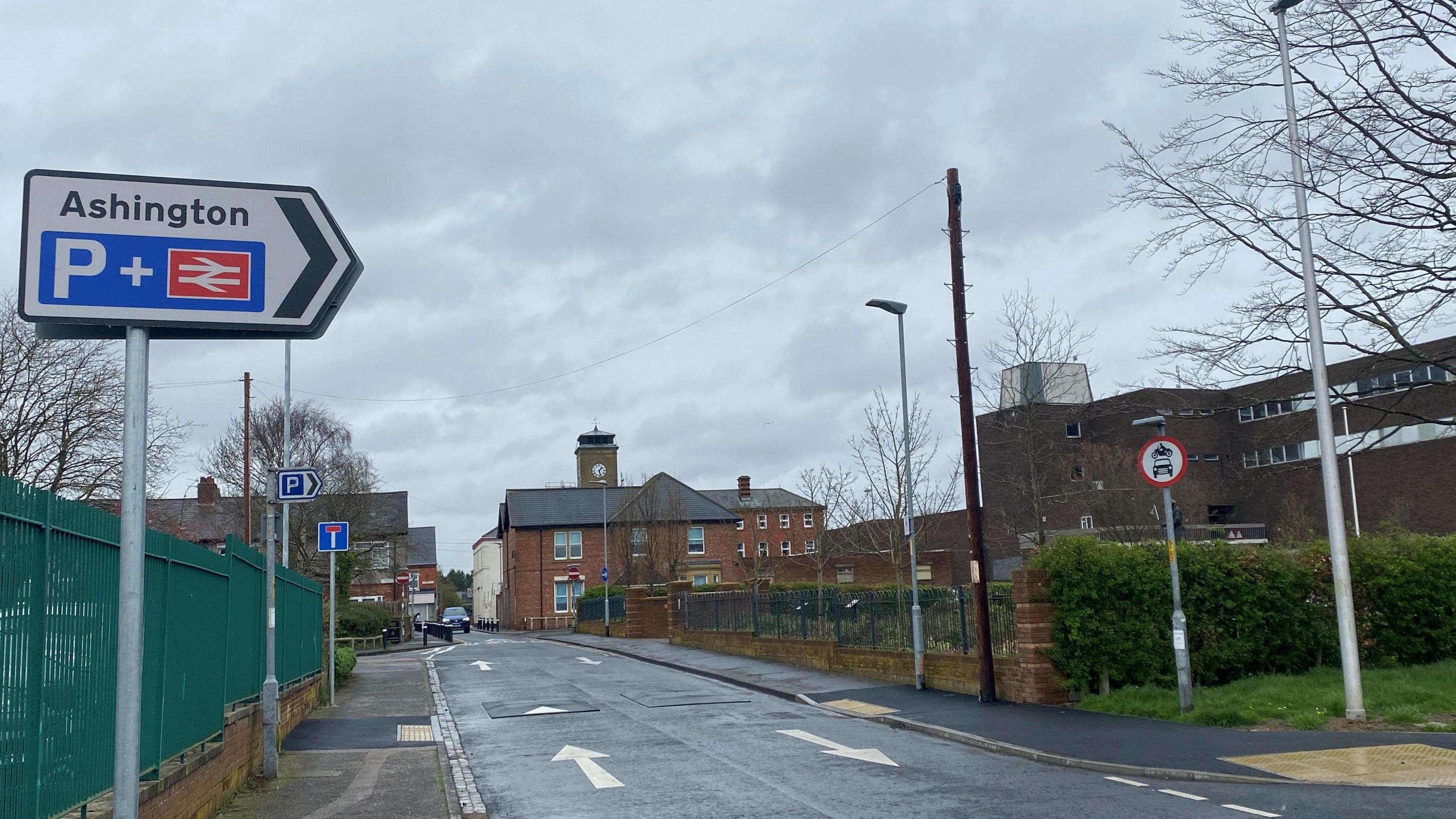 A red sign for Ashington station overlooking a road without traffic