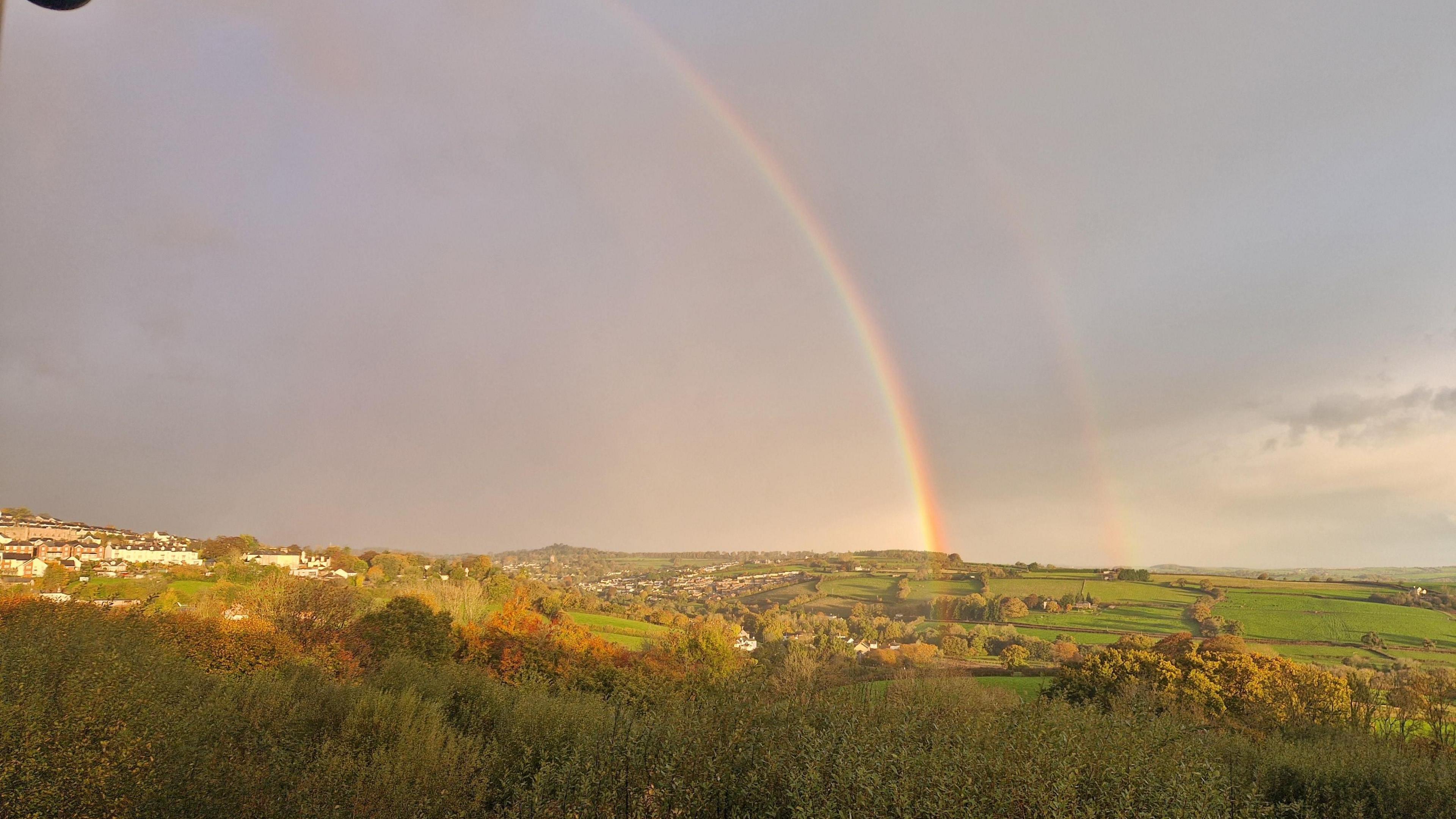 A double rainbow. The one on the left is brighter than the one on the right. The sky is grey and filled with clouds. There are green fields and green trees. 