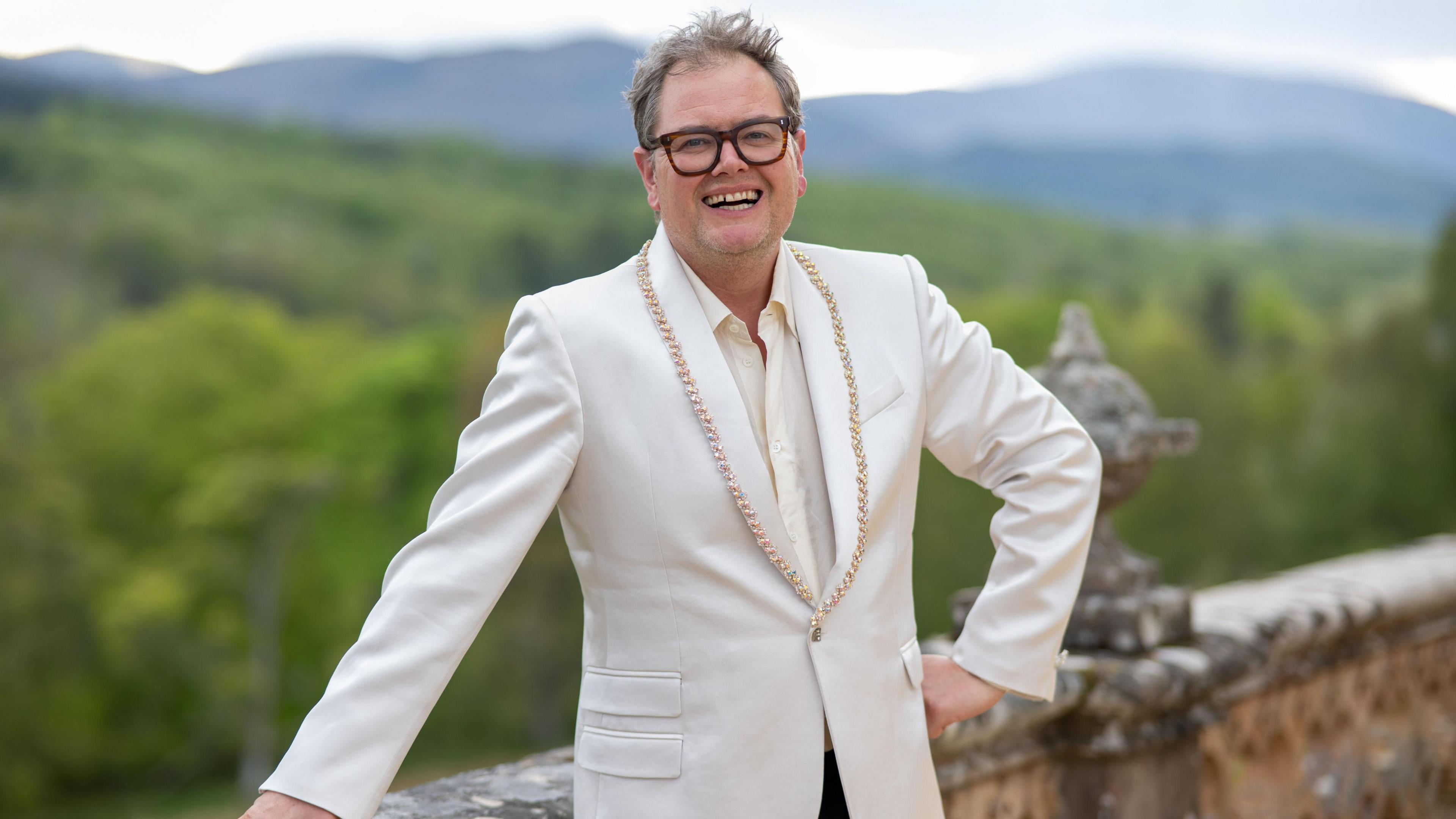 Alan Carr. He is stood on a balcony overlooking the Scottish Highlands. He is wearing a white suit and glasses with thick black frames. He has one hand on his hip and the other on the balcony. He is looking at the camera and smiling.