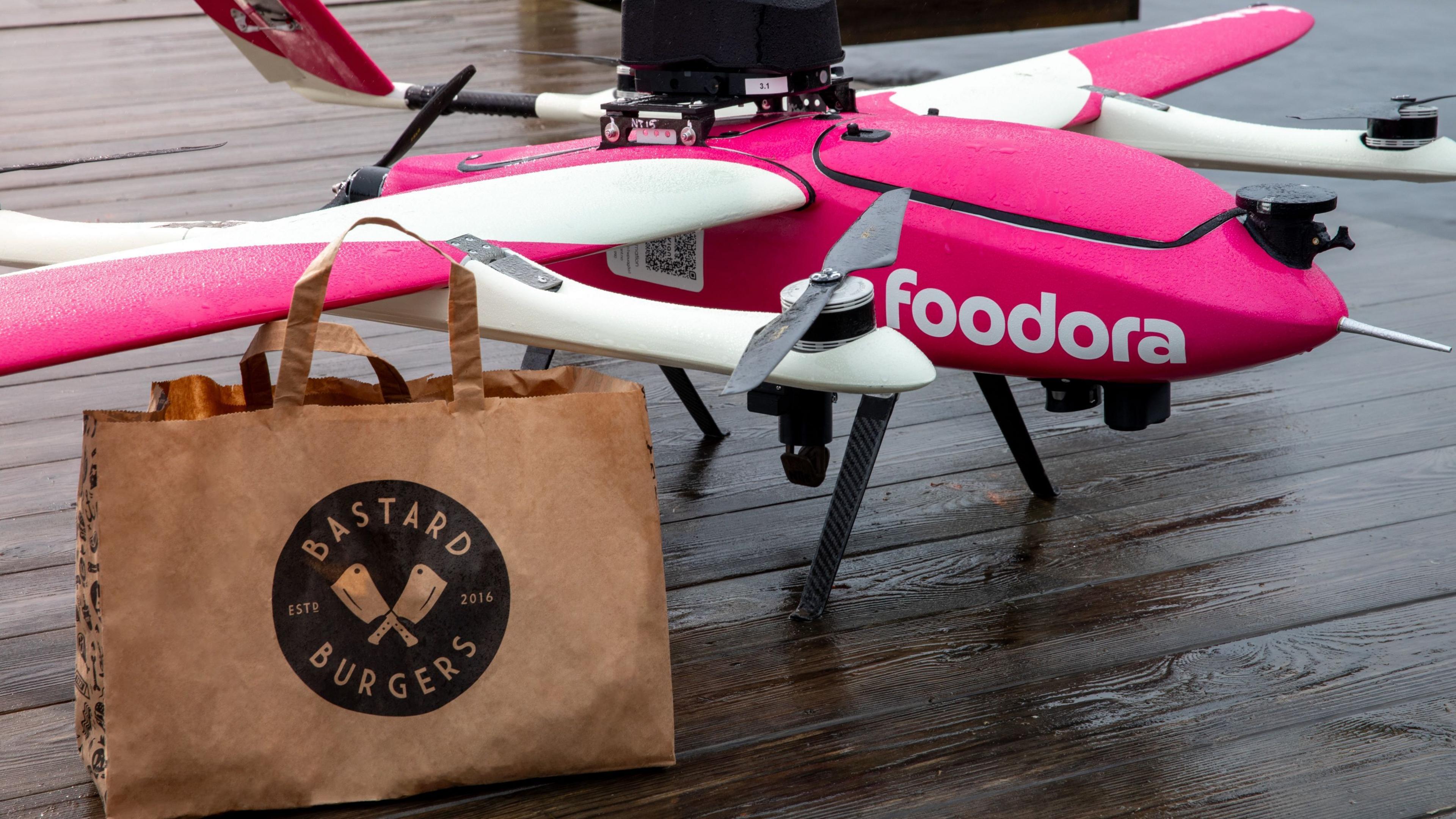The foodora drone is pink and white with rotors on mounts attached to the wings - a third propeller is on the back. In the foreground is a brown paper bag from Bastard Burgers.