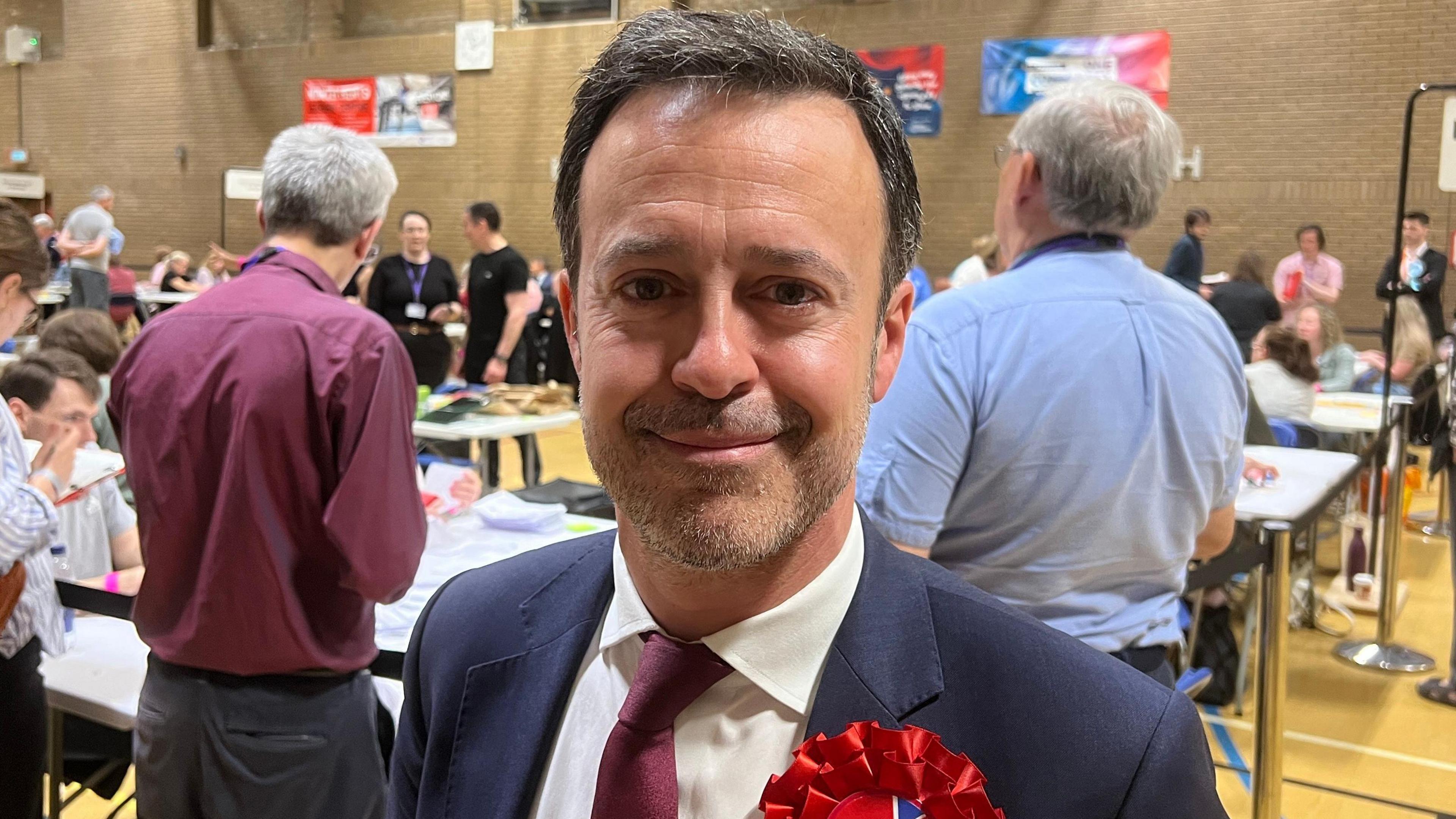 Kevin Bonavia with short dark hair, smiling at the camera at an election count. He has a blue jacket, white shirt and red tie. A red rosette is pinned to his jacket. There are counting tables behind around which people are standing.