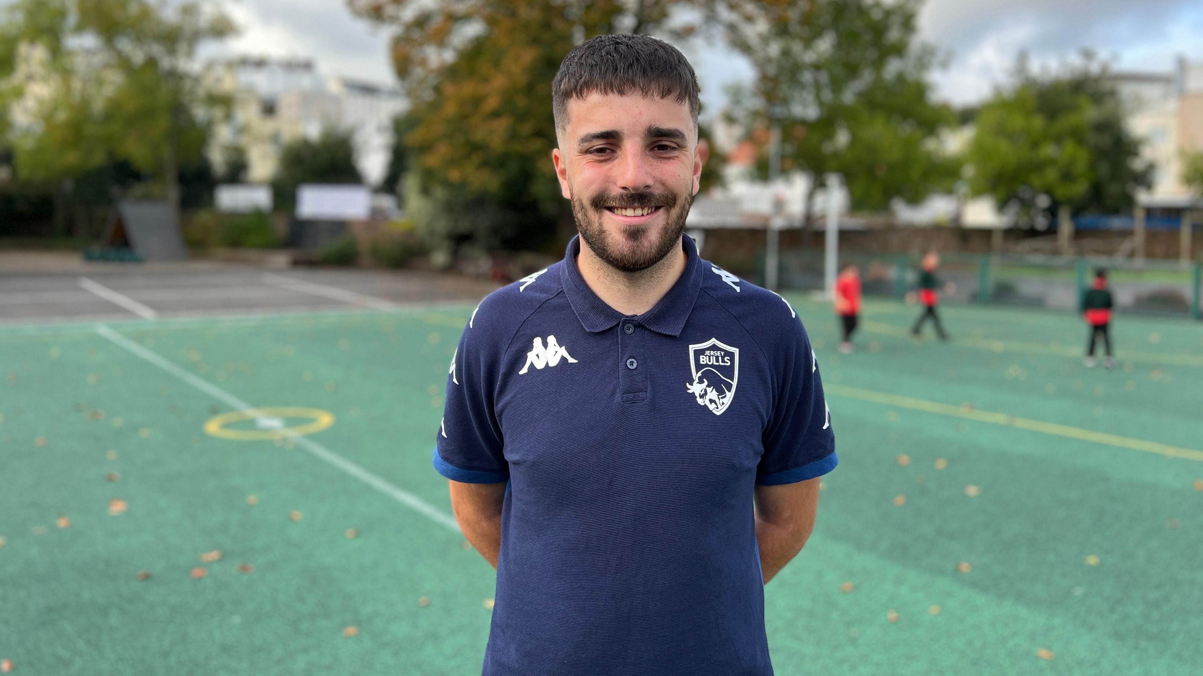 Michael smiles at the camera and his hands are behind this back. He is wearing a navy blue polo top with a white Jersey Bulls logo on his chest. Three children are playing football behind him. Michael has dark hair which is shaved on the sides and he has a stubbly beard.