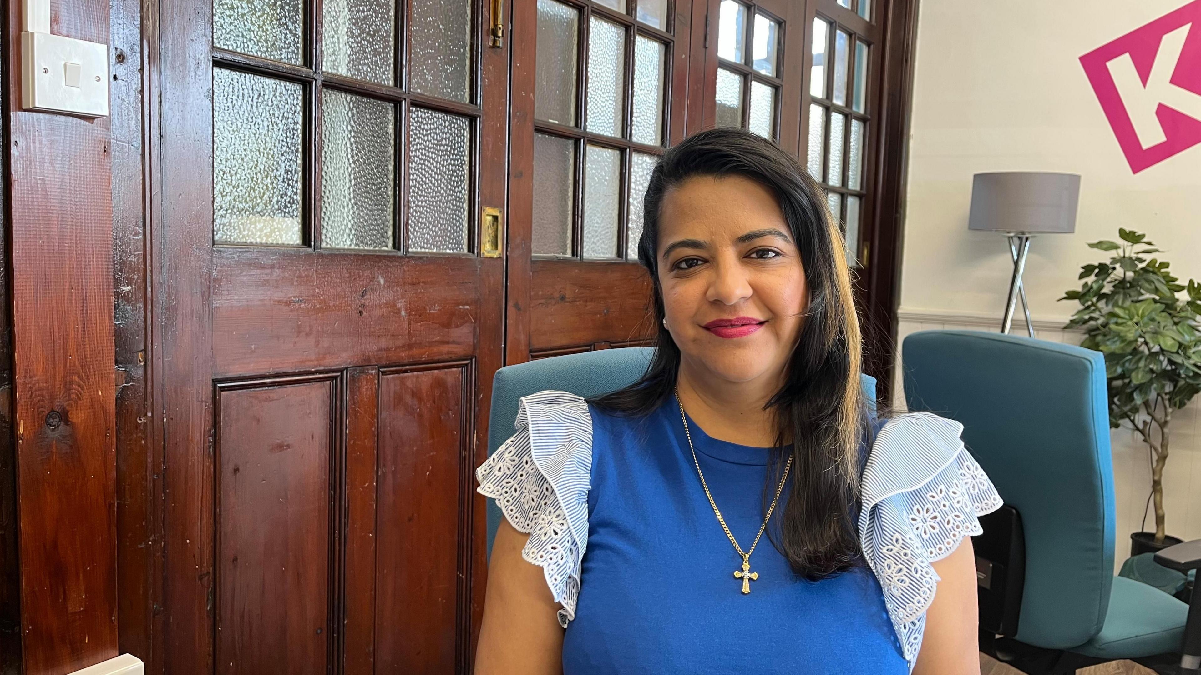Melisha Pillay looks directly at the camera as she is photographed sitting in a room with a dark wooden door behind her. She is wearing a blue top with stripey, ruffled sleeves. She has a long gold chain around her neck with a cross hanging from it. She has dark hair.