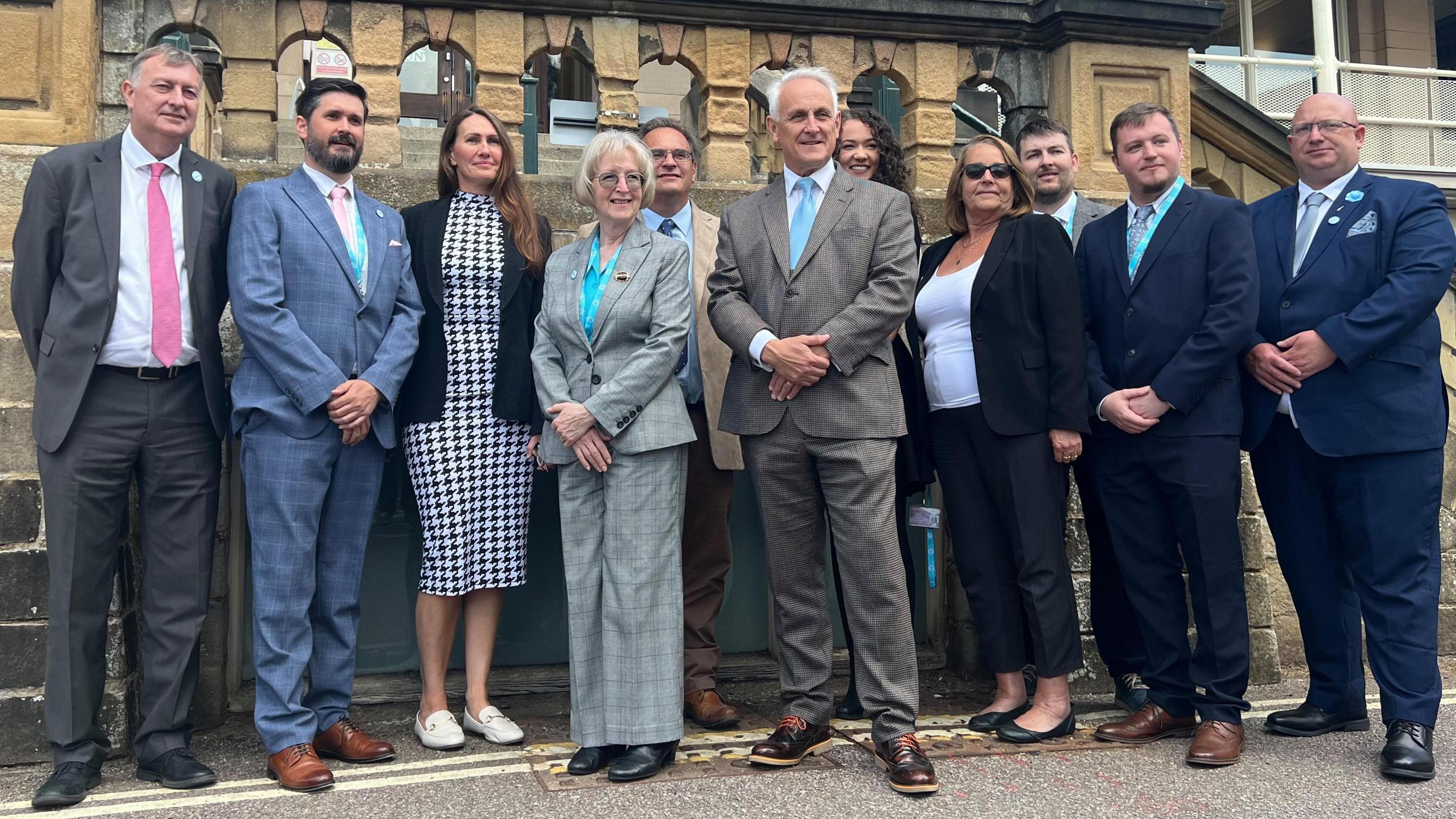 11 people in suits stood infront of grey stone building