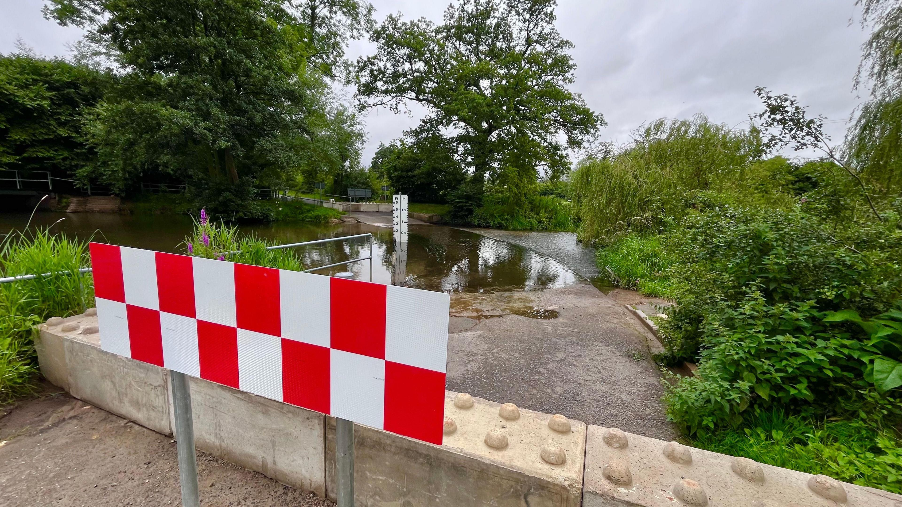 A view of the concrete barriers at the edge of Shotesham ford. It shows the barriers placed across the entire length of the road while the ford can be seen in the distance.