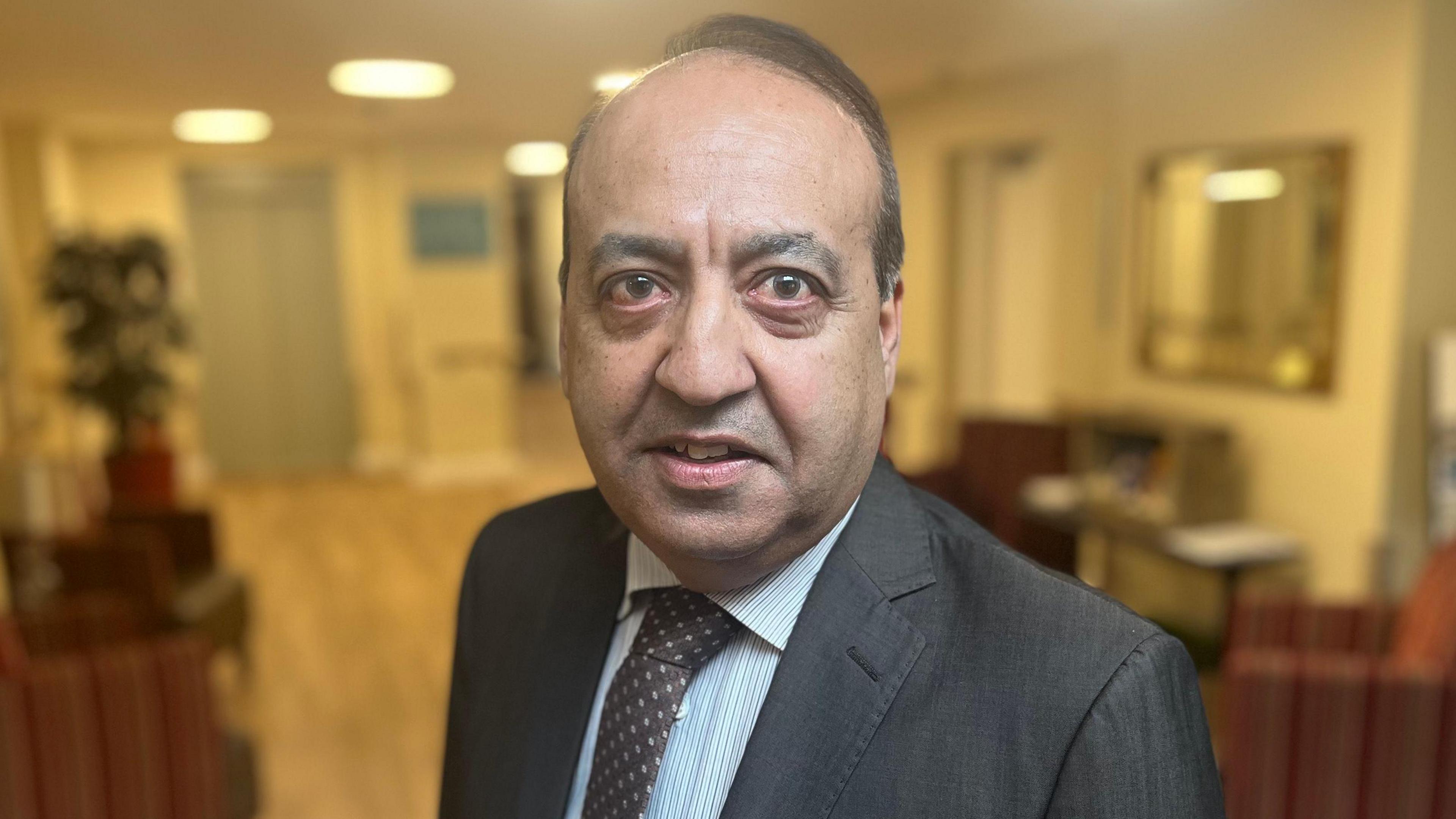 Zuffar Haq wearing a dark grey suit, white and blue striped shirt and dark tie.