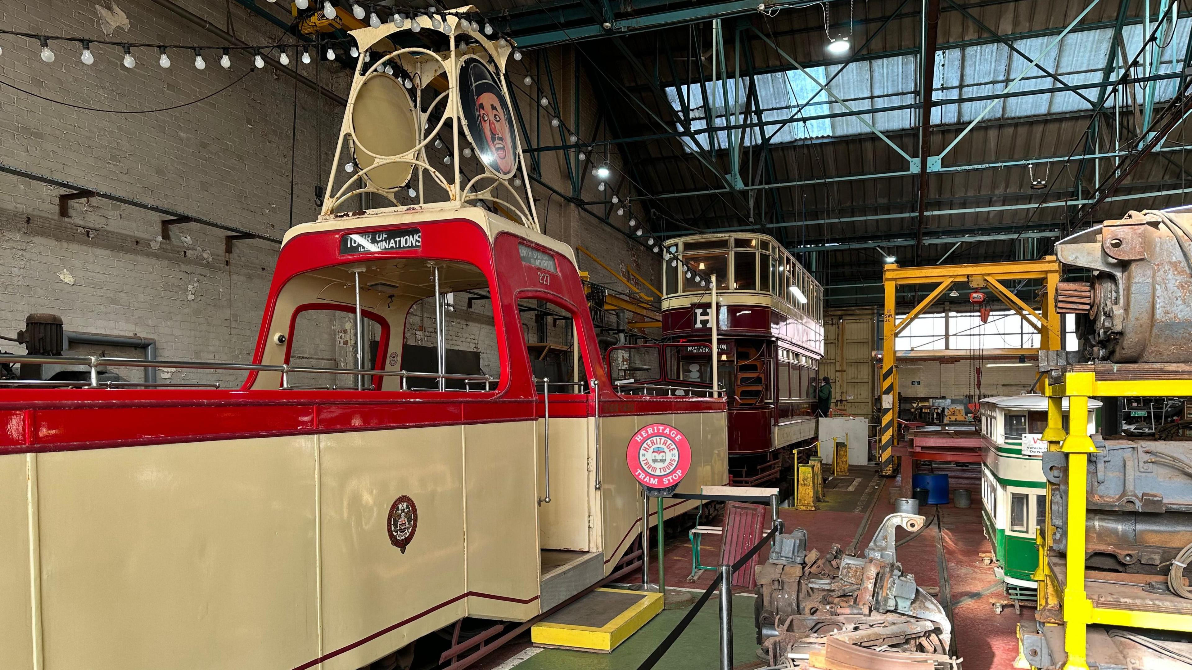 The red and cream Charlie Cairoli boat tram is positioned in the warehouse in front of a maroon-coloured double decker tram. 