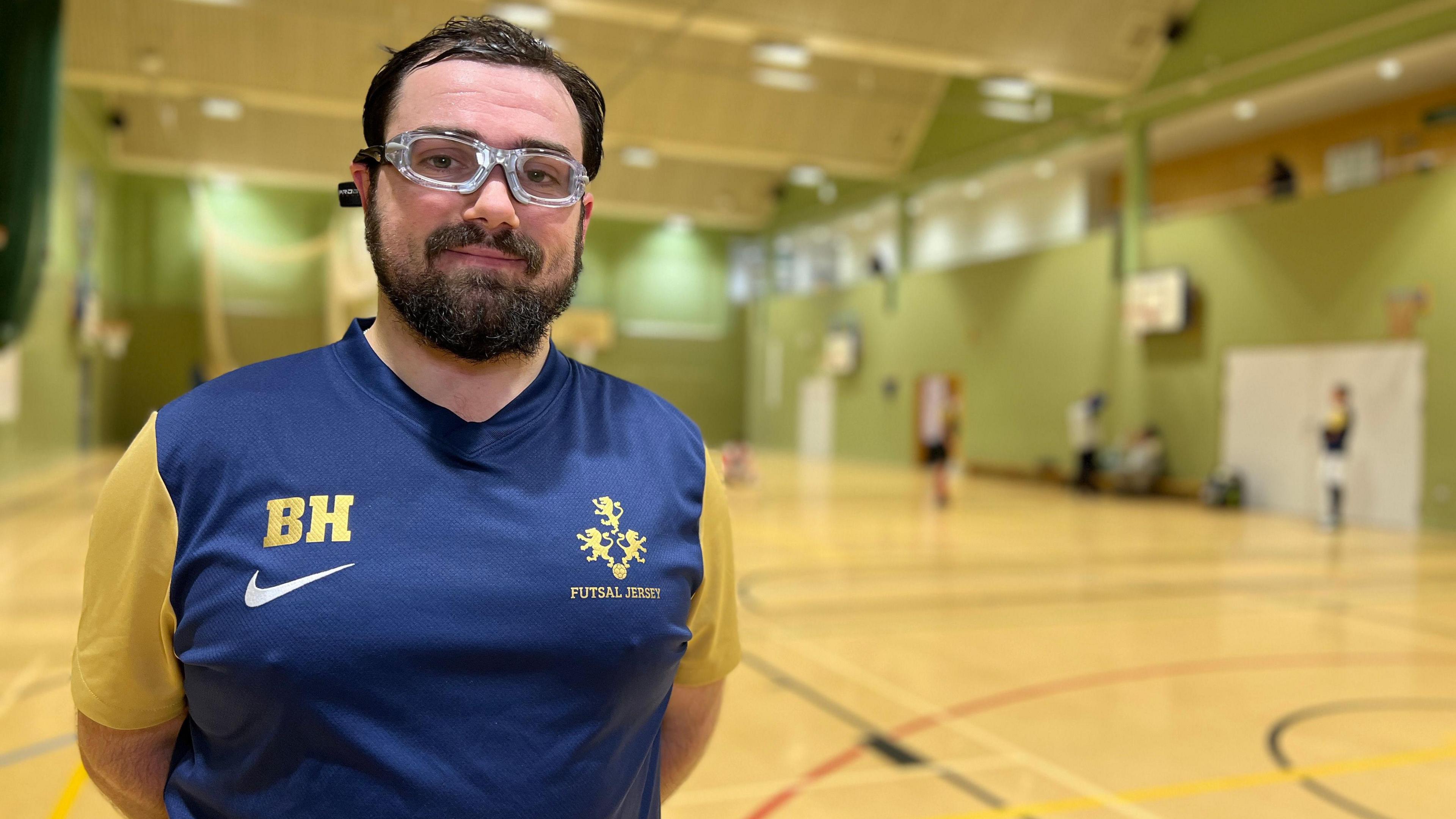 Bernardo smiles at the camera as people play futsal in the sports hall behind him. He is wearing protective goggles and a blue football shirt with yellow sleeves. He short dark hair and a dark beard.