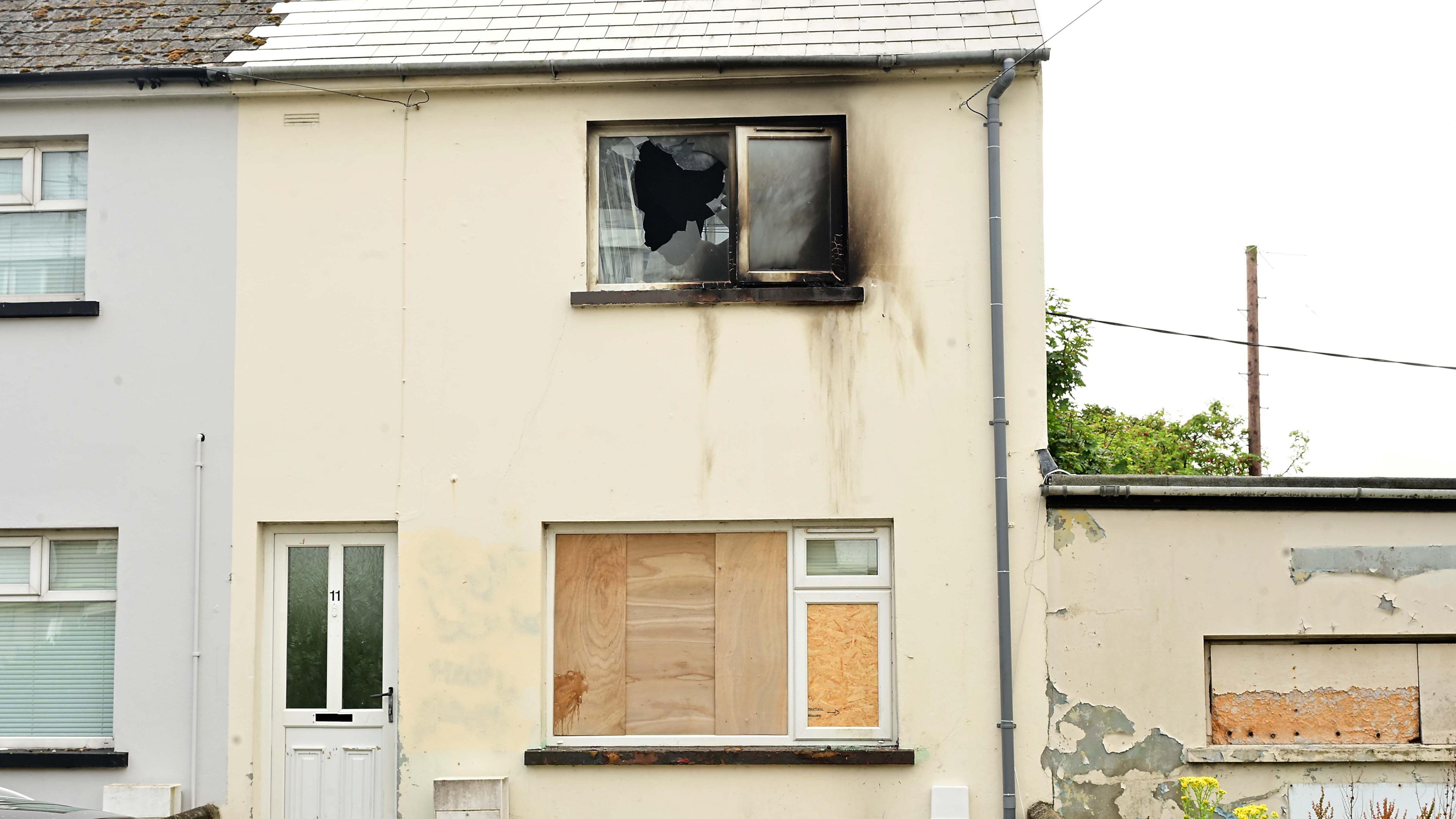 A cream coloured house with boarded windows and an upstairs window that is smashed and burned. There are burn marks down the wall.